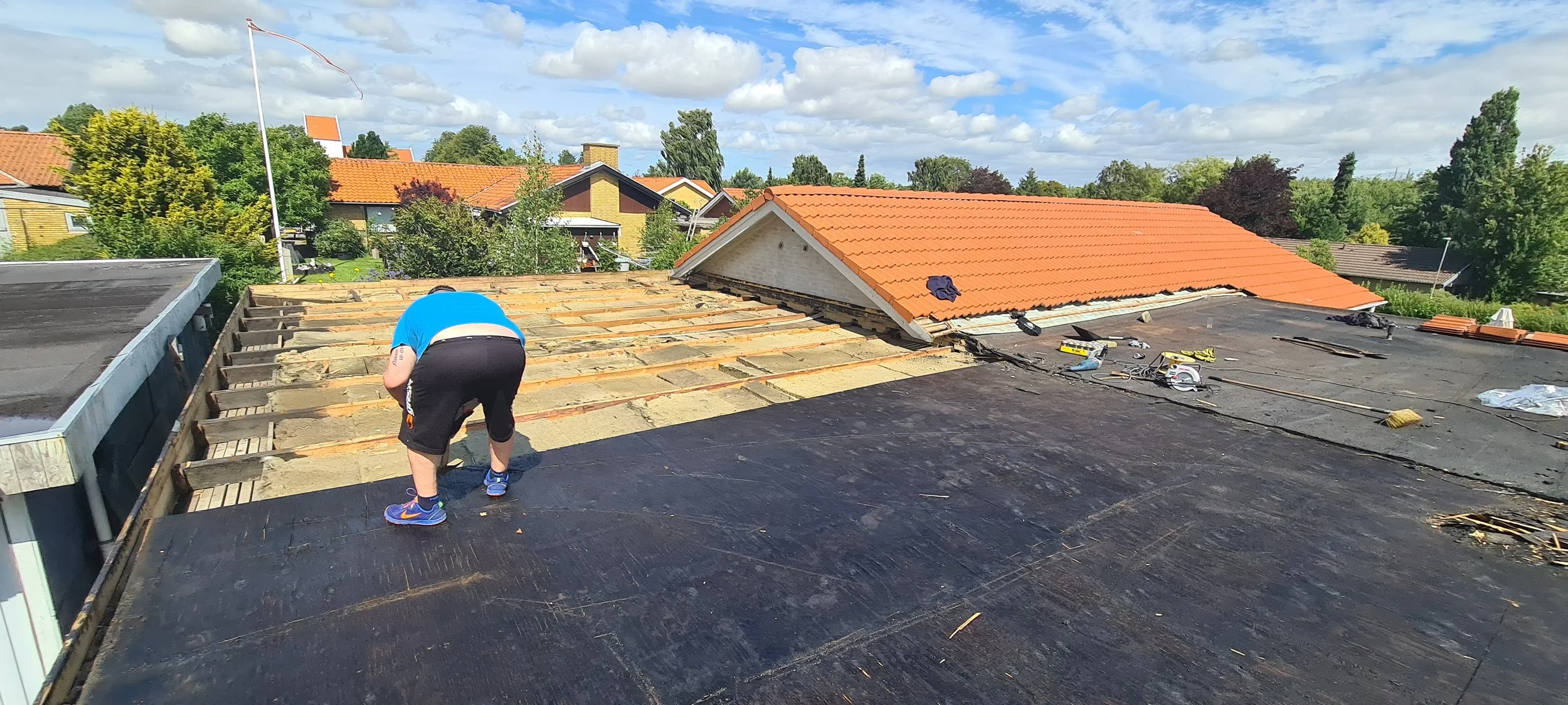 A person working on the roof of a house, removing old shingles and preparing for new roofing material. The sky is partly cloudy and nearby houses with red-tiled roofs are visible.