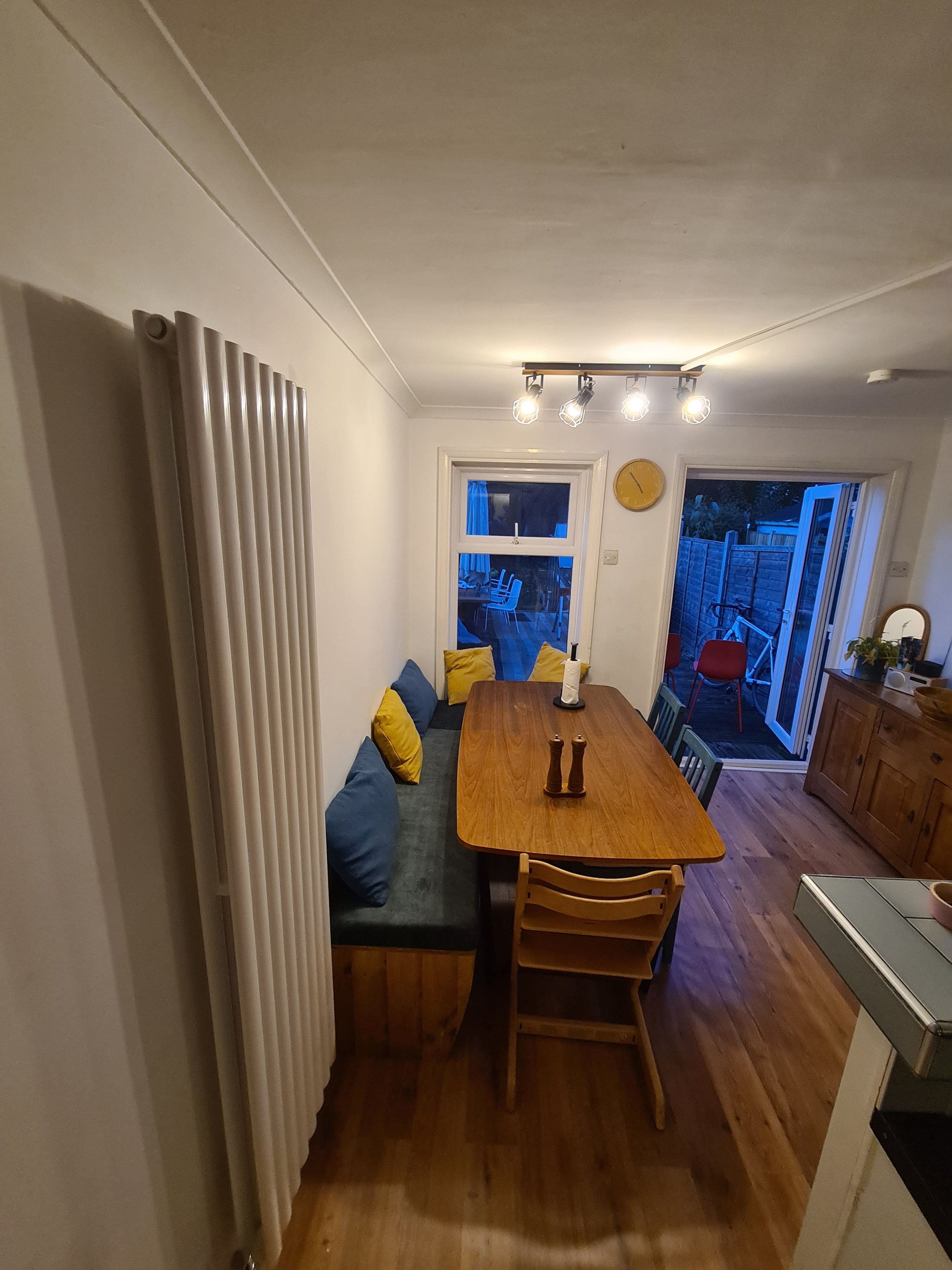 Dining room with wooden table, yellow and blue cushions on bench, open sliding door to outdoor patio, and window with blue evening sky outside.
