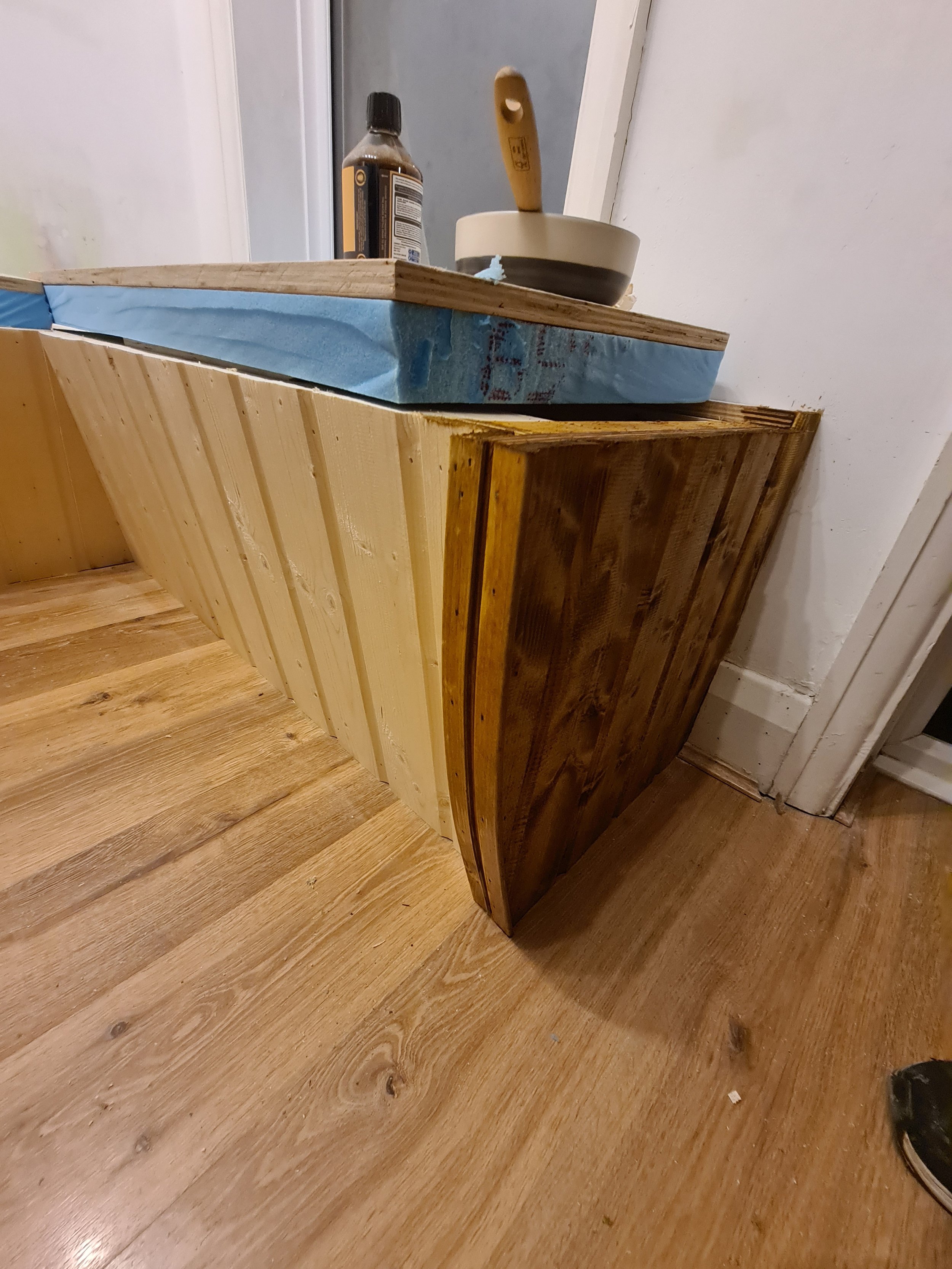 A corner of a room with a wooden bench or table, and a black bottle, paintbrush, and white bowl on top. The furniture is made of different types of wood, with some parts varnished and others unfinished.