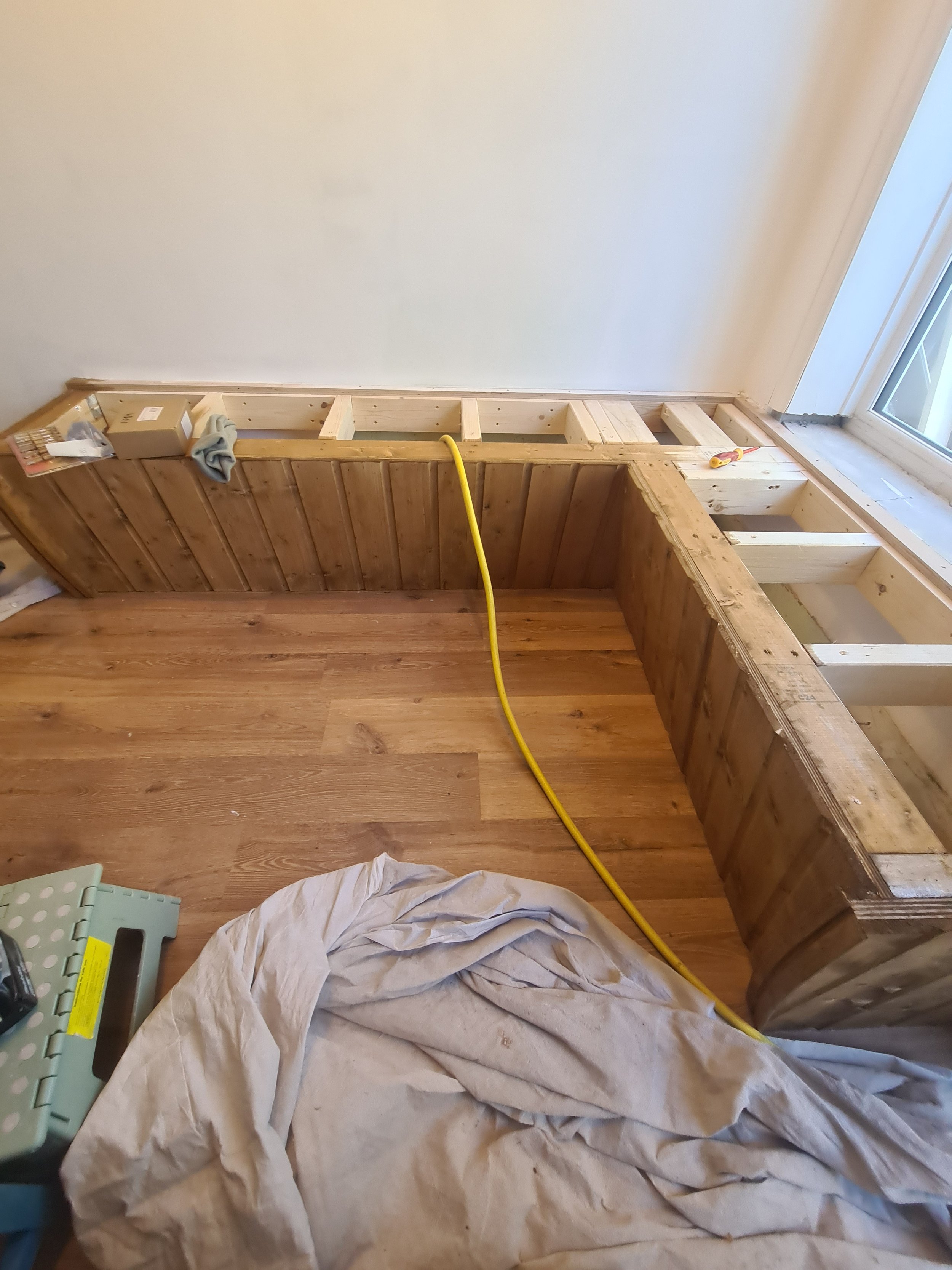Room under renovation with a wooden framework, tools, a yellow extension cord, and a window with sunlight.