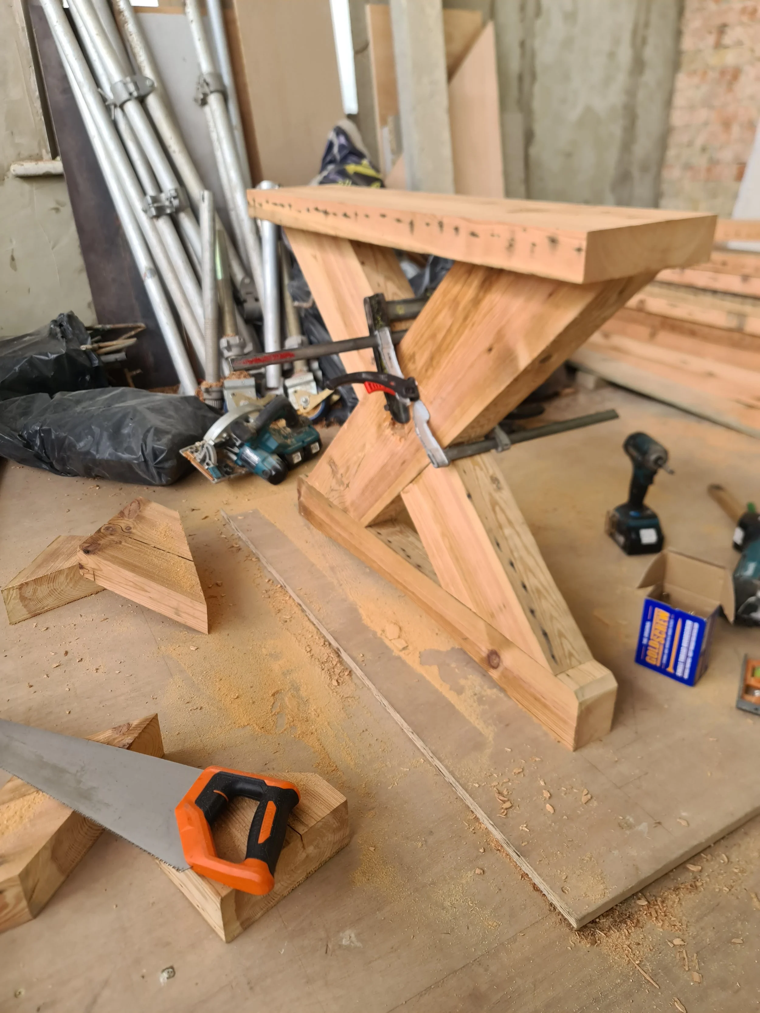 A woodworking project in progress, featuring a wooden sawhorse or stand held together with clamps, with other tools such as a hand saw, drill, and various pieces of wood scattered on the work surface at a construction site.