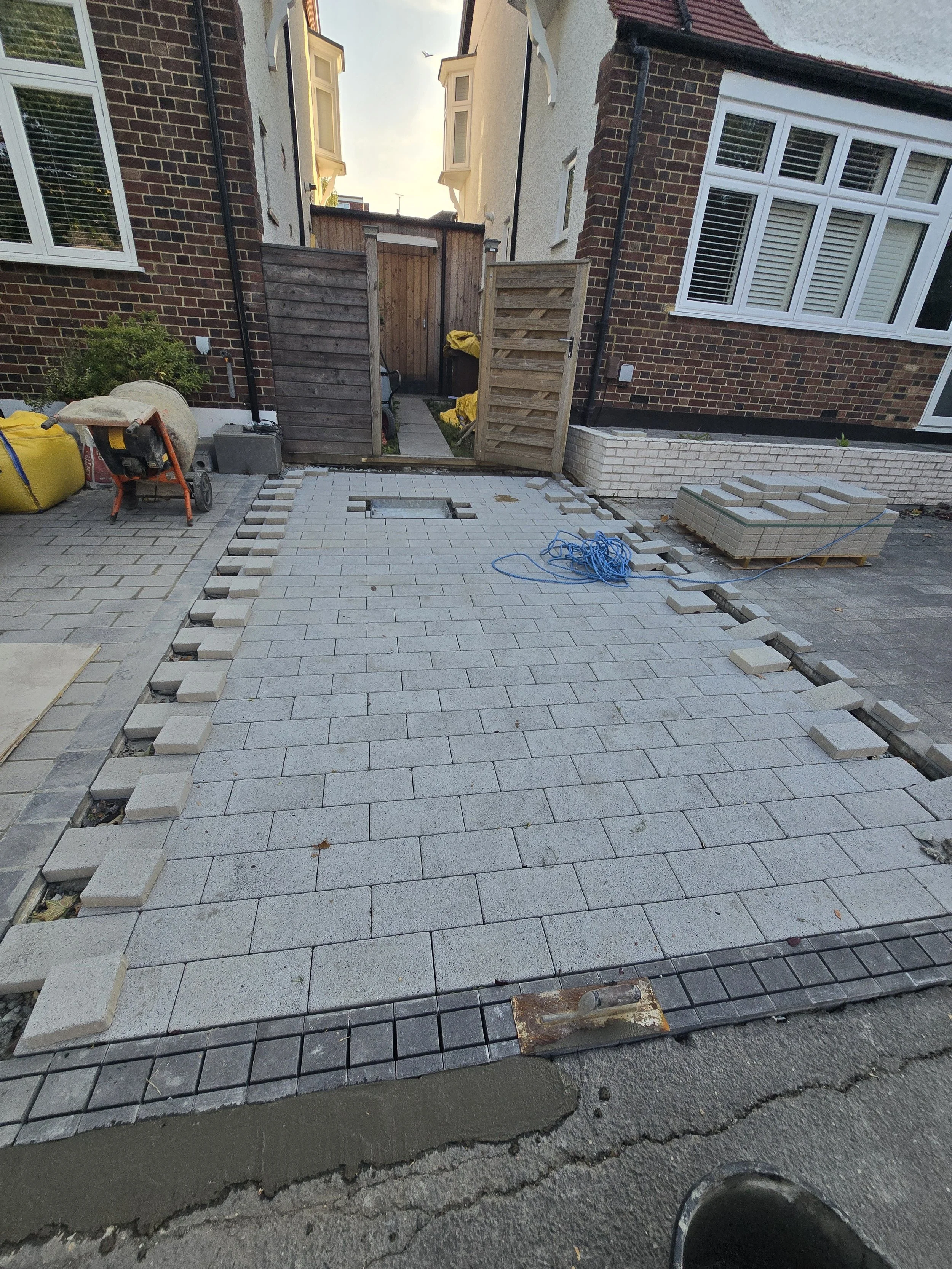 A driveway under construction with gray paving stones, some stacked on the side, and a partially completed border. There's a wheelbarrow, yellow bags, and construction tools nearby, with a wooden gate at the back.