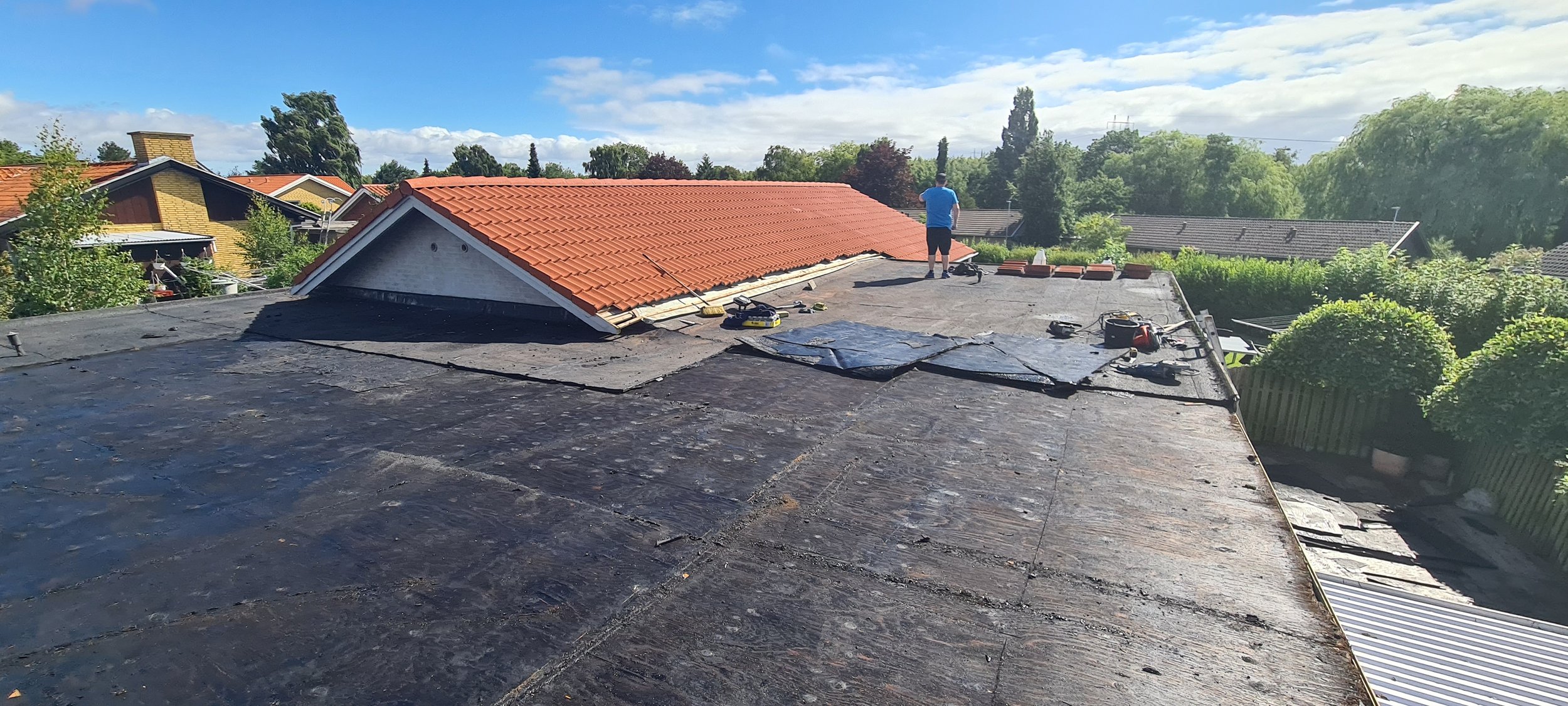 A person on a roof working with roofing tools and materials, with neighboring houses and green trees in the background under a partly cloudy sky.
