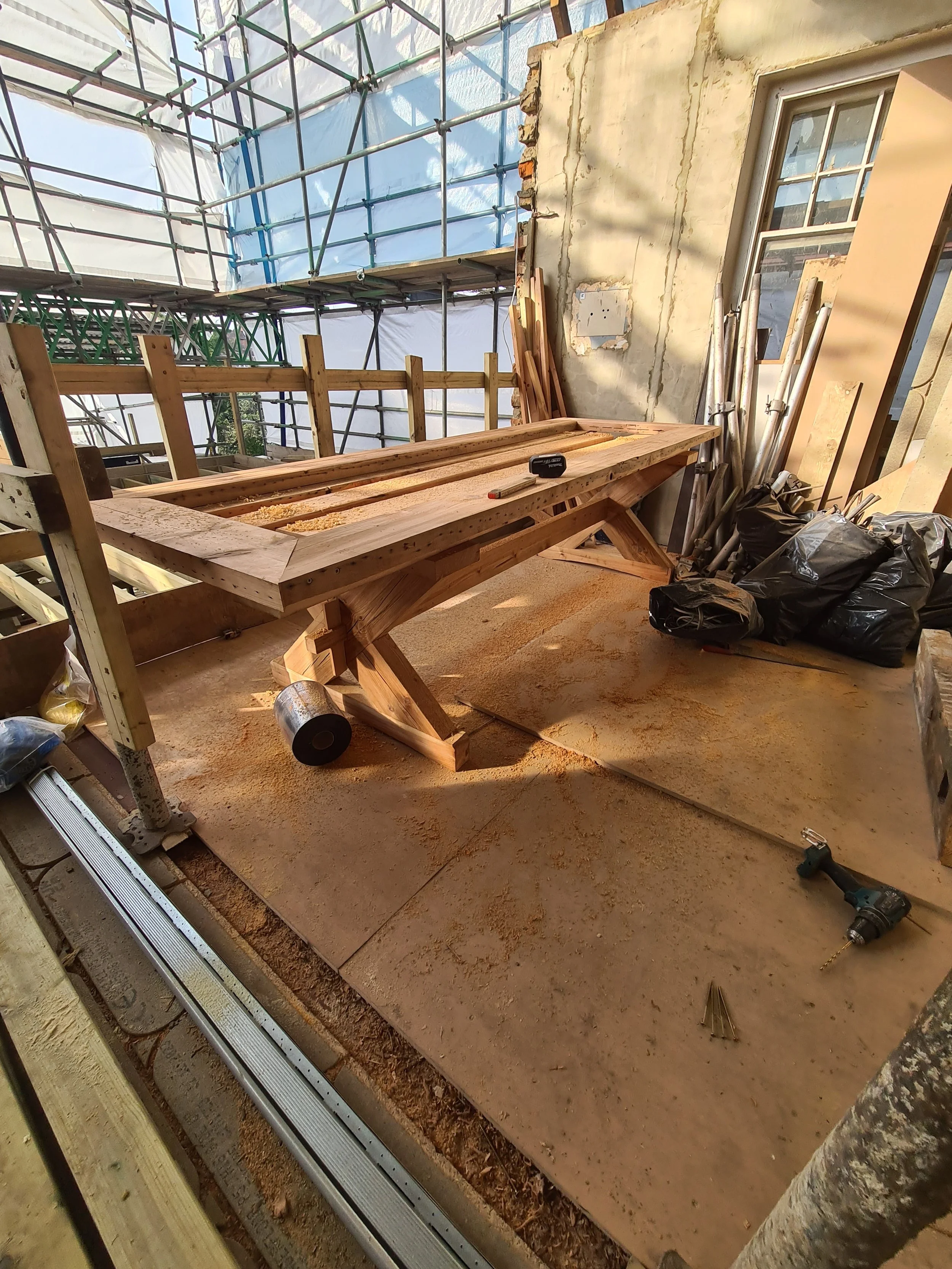 Construction site showing a wooden workbench, power drill, sawdust, and construction materials inside a partially built structure with scaffolding outside.