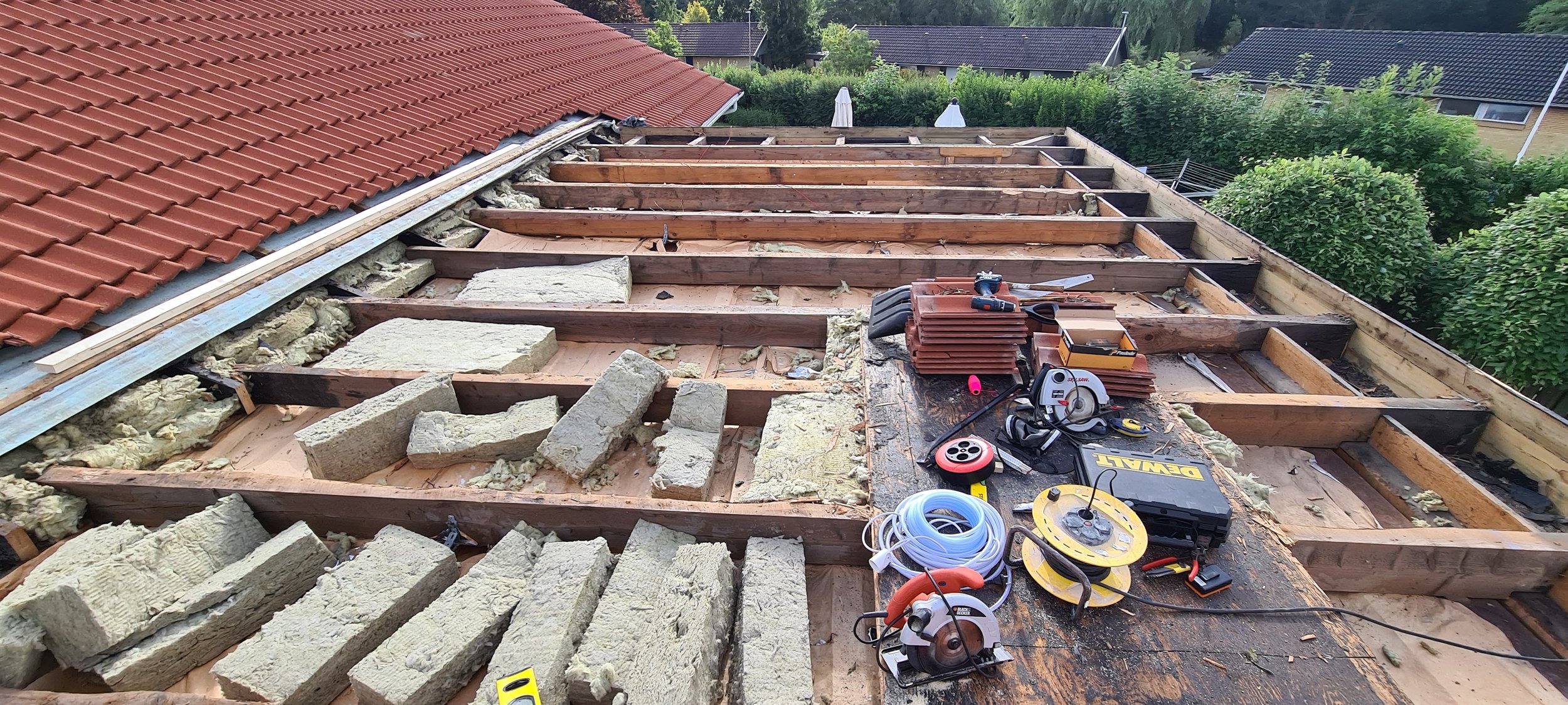 Construction site on a house roof with tools and materials, including insulation, wooden beams, and power tools, in a residential neighborhood.