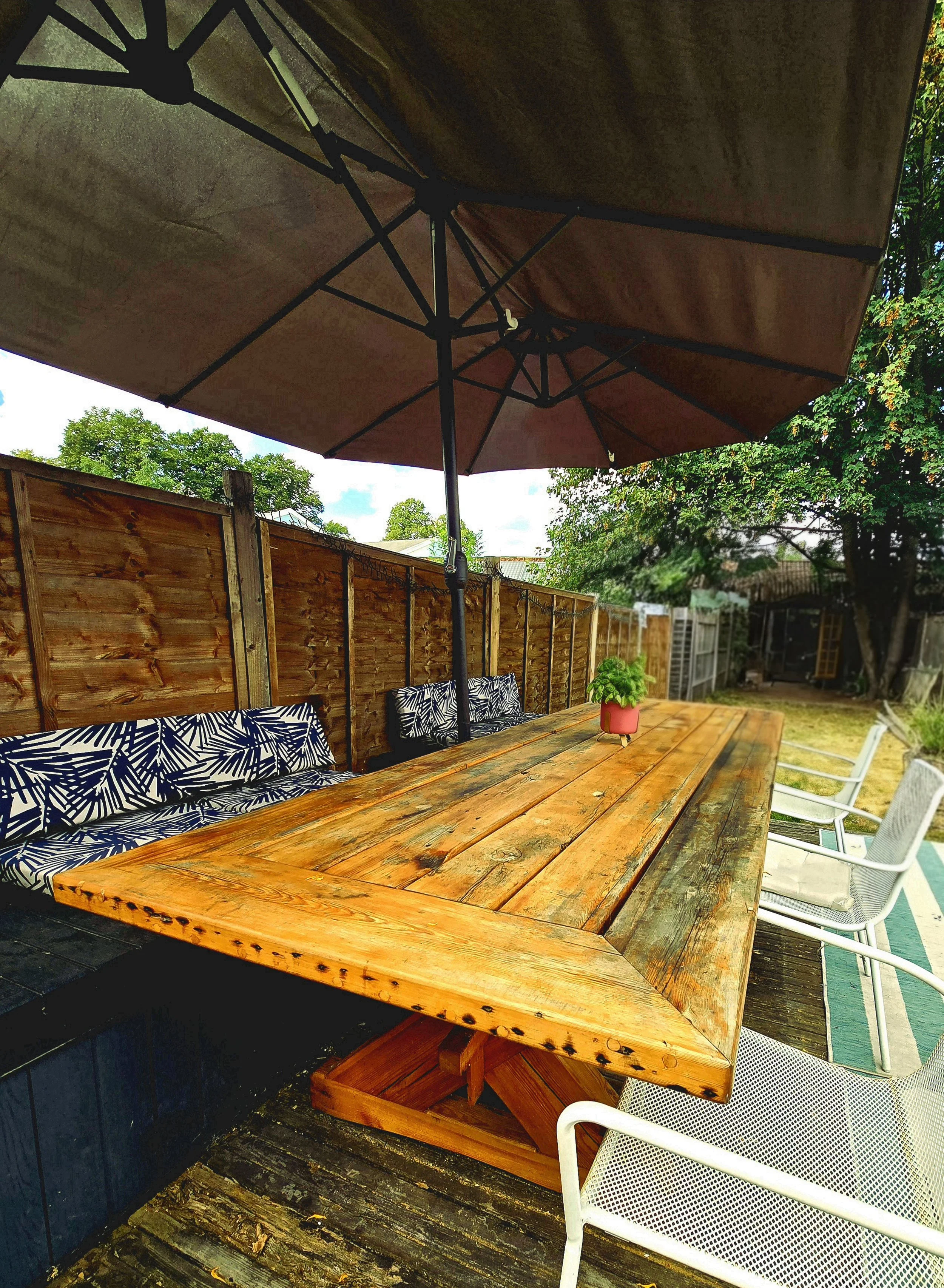 Outdoor patio setup with a large wooden dining table, white chairs, a large umbrella, a potted plant, and a wooden privacy fence. There are trees and a clear sky in the background.