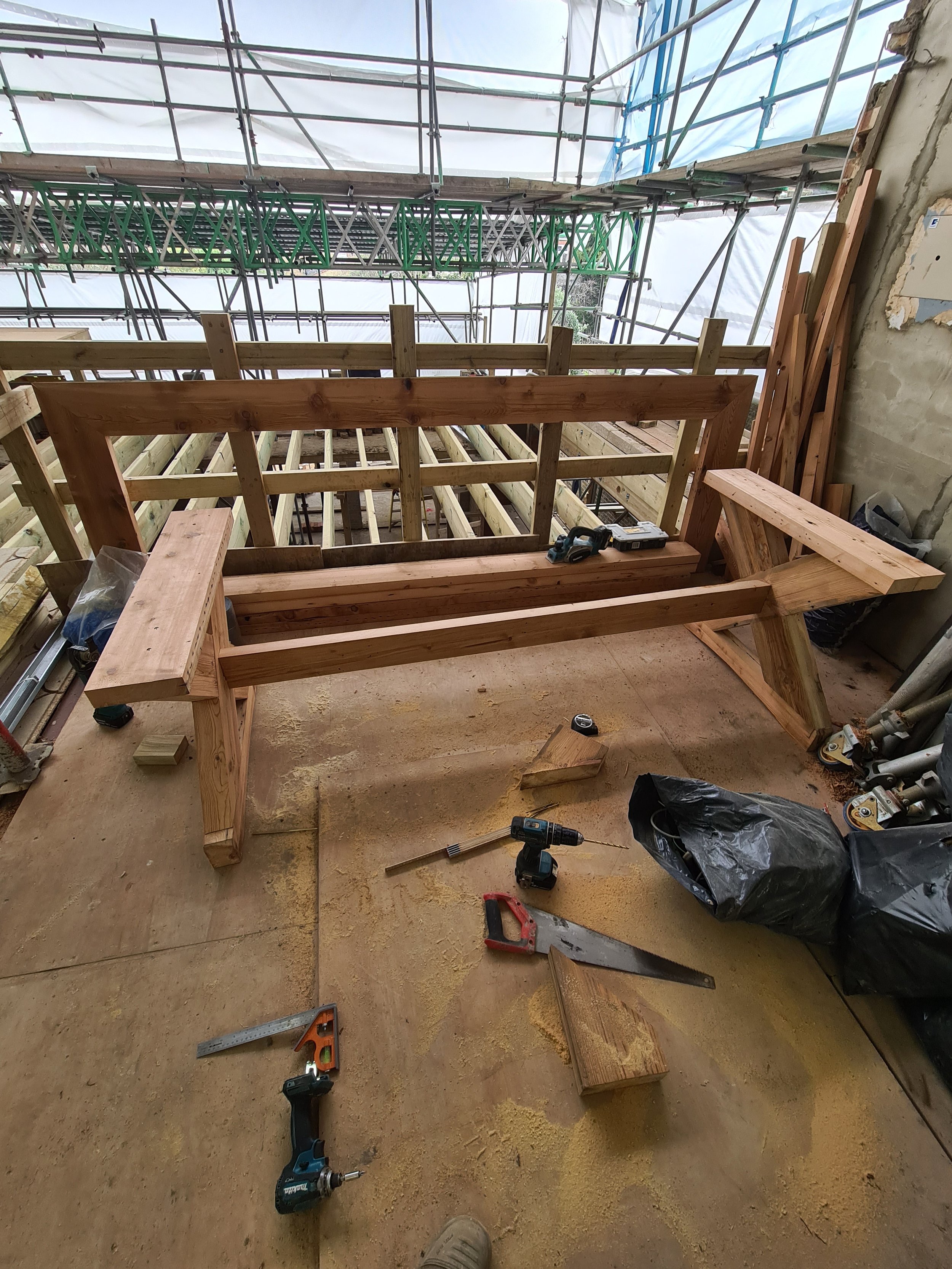 Construction site with wooden framework and tools. Scaffolding and safety netting are visible in the background, as well as construction materials and equipment on the floor.