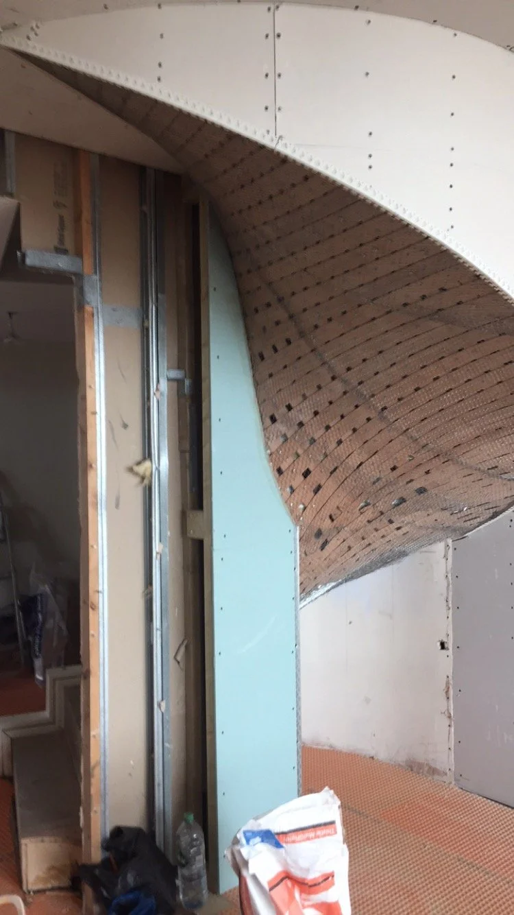 Construction site showing part of an arched ceiling with wooden slats and drywall, exposed framing, and construction materials.