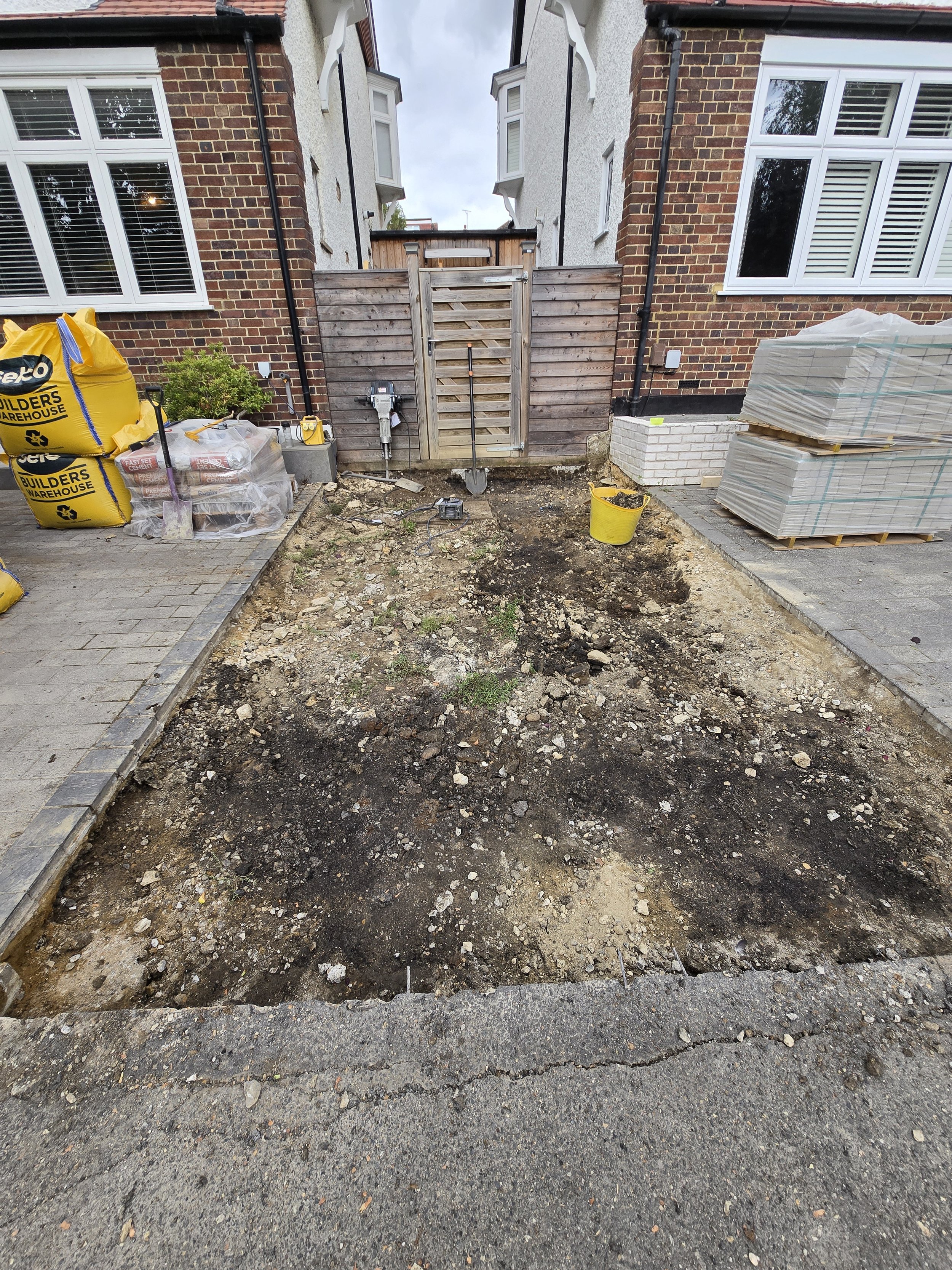 A backyard with an area of soil being prepared for landscaping or gardening, surrounded by paved pathways, with construction supplies and tools, a wooden gate at the backyard's fence, and houses with brick walls and windows in the background.