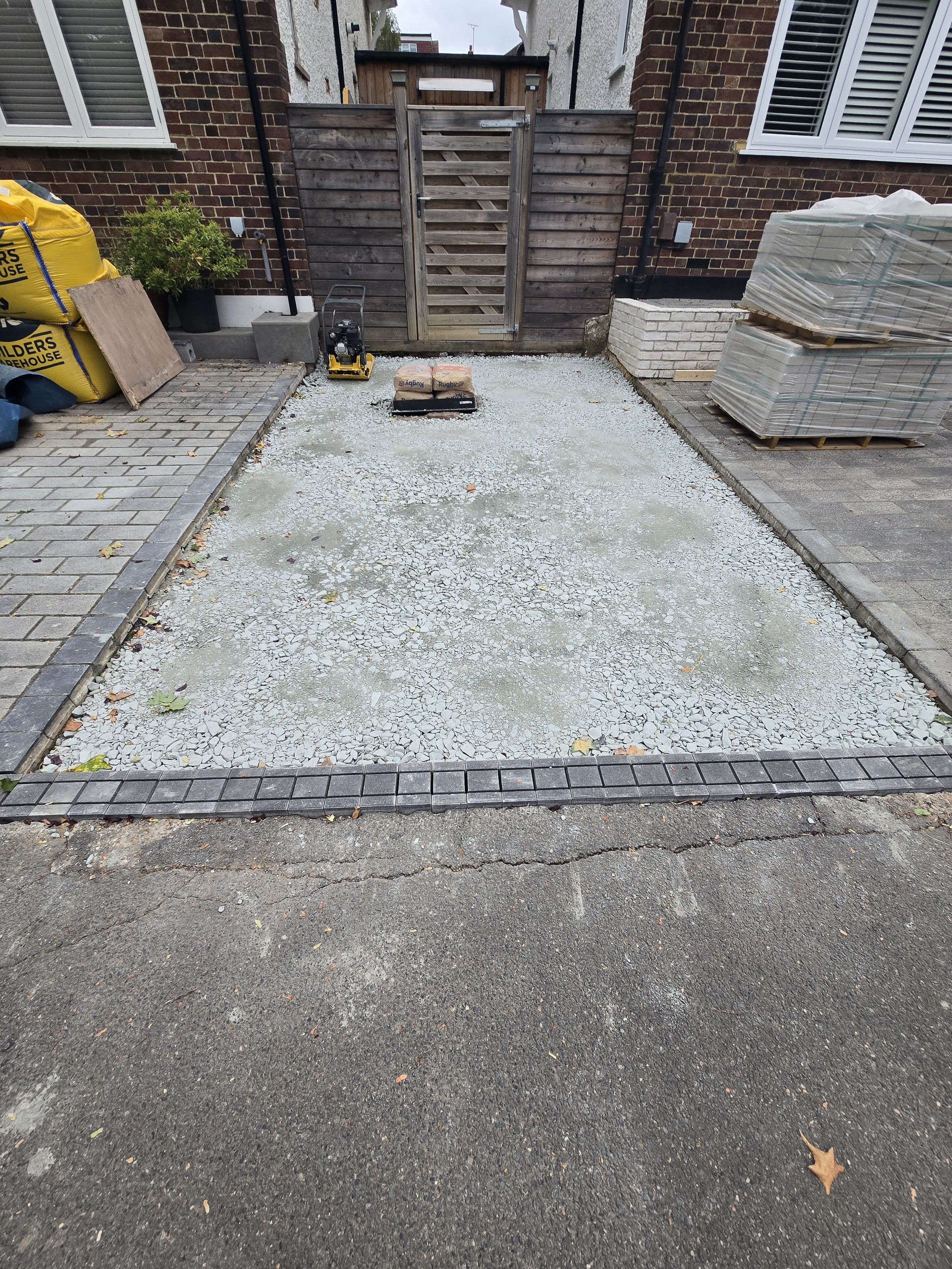 A driveway under construction with gravel spread out, surrounded by paved borders, with construction materials and tools present, leading to a gate at the back. The area is enclosed by brick and white-sided houses.