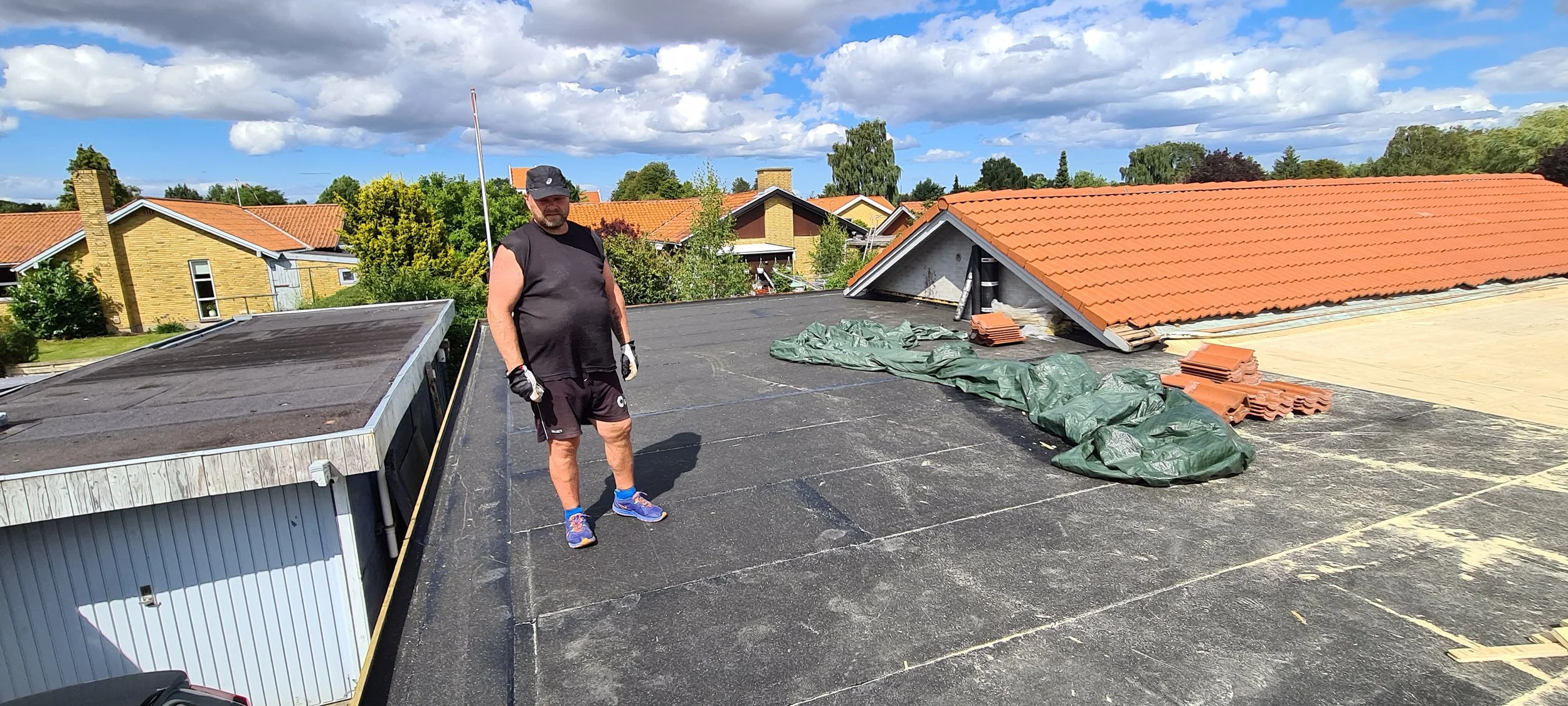 Man standing on a flat rooftop during a roofing project with materials and tiles nearby, under a partly cloudy sky.