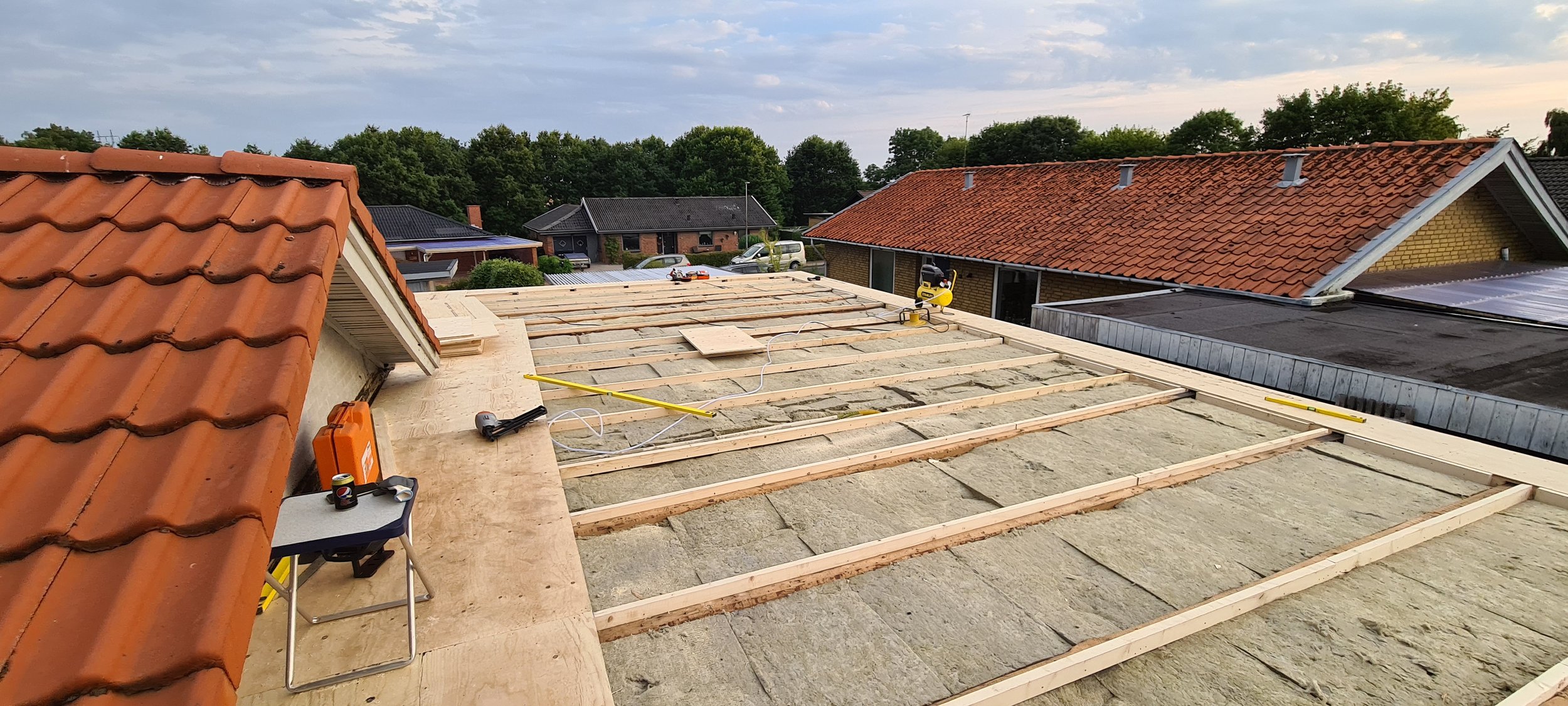 Roof under construction with wooden framework, tools, and materials, neighboring houses with tiled roofs, trees in the background, and cloudy sky.