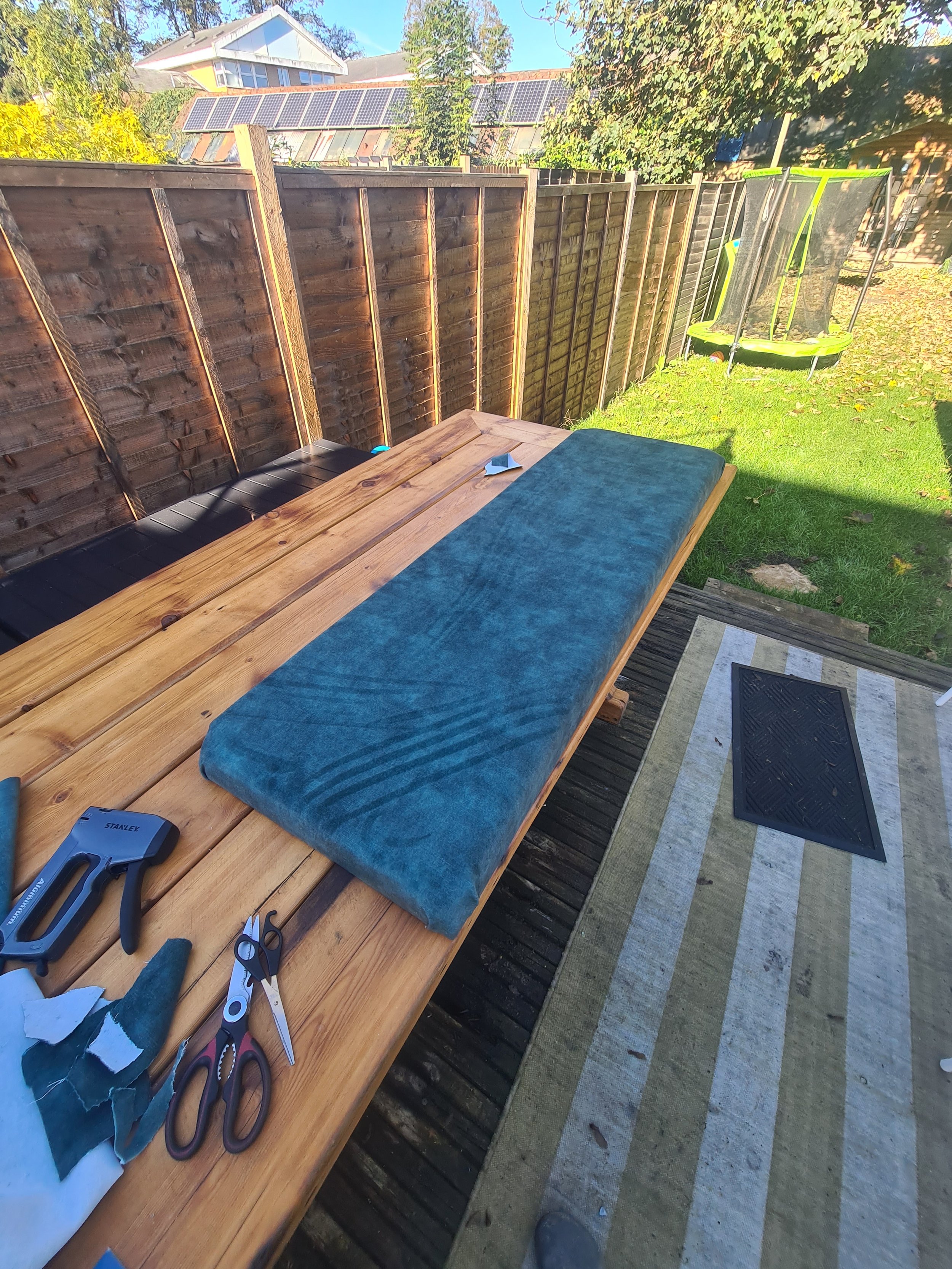 Outdoor workspace with a wooden table, tools, and a foam cushion. In the background, a fenced backyard with a trampoline and a house with solar panels.