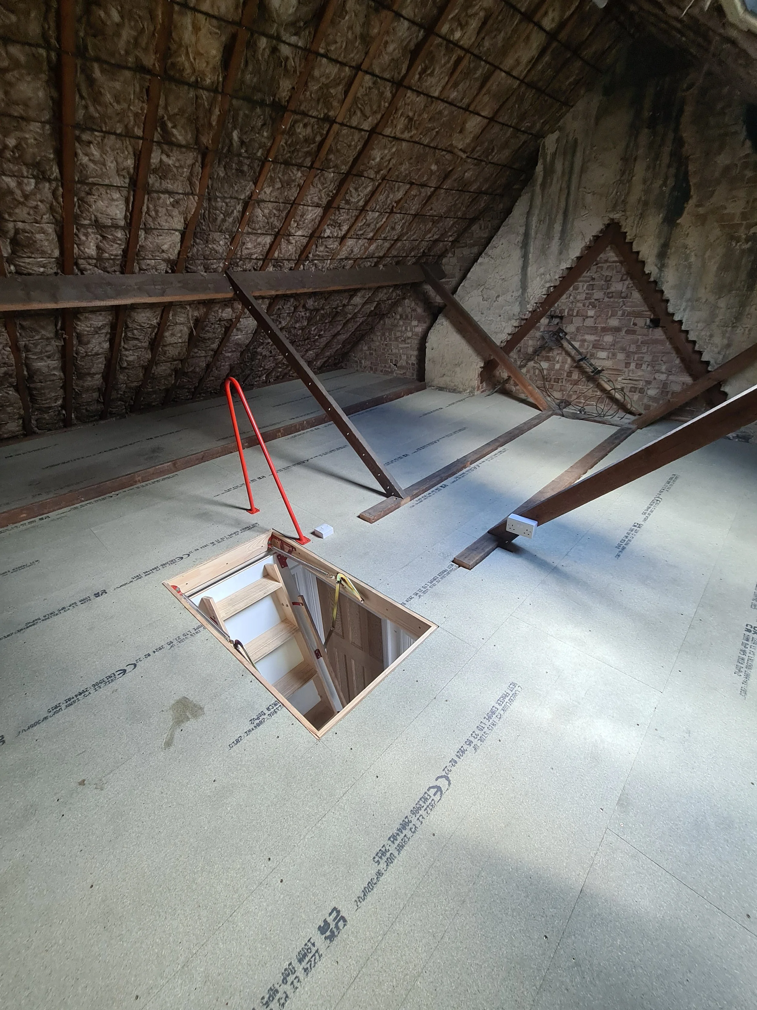 Attic with exposed roof framing, insulation, and a small hatch in the floor.