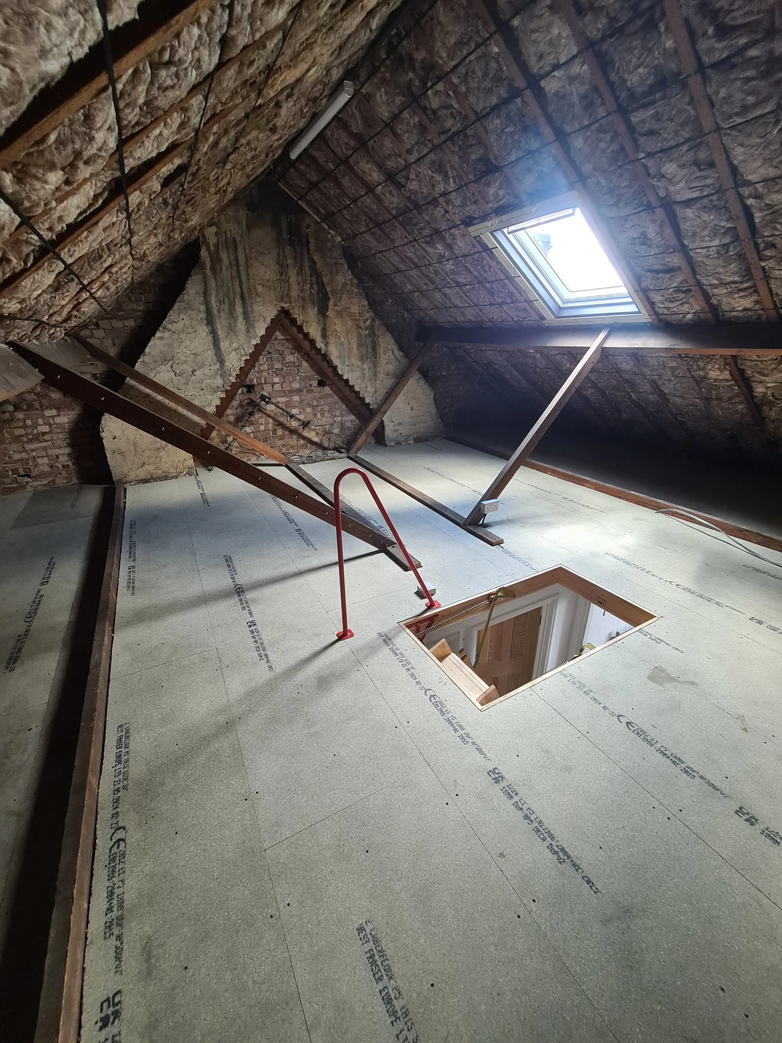 Attic space under renovation with insulation and drywall removed, showing exposed brick walls, a skylight window, and an opening in the floor.