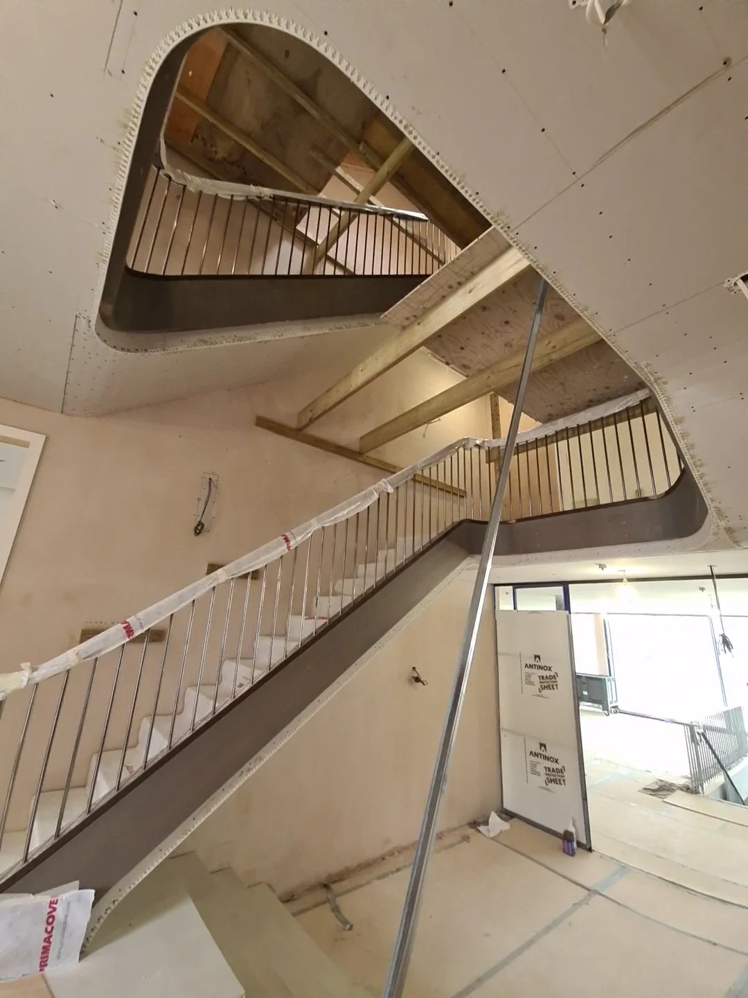 Interior view of a building under construction showing a staircase with metal railings, drywall installation on the ceiling, and exposed wooden beams and partial flooring.