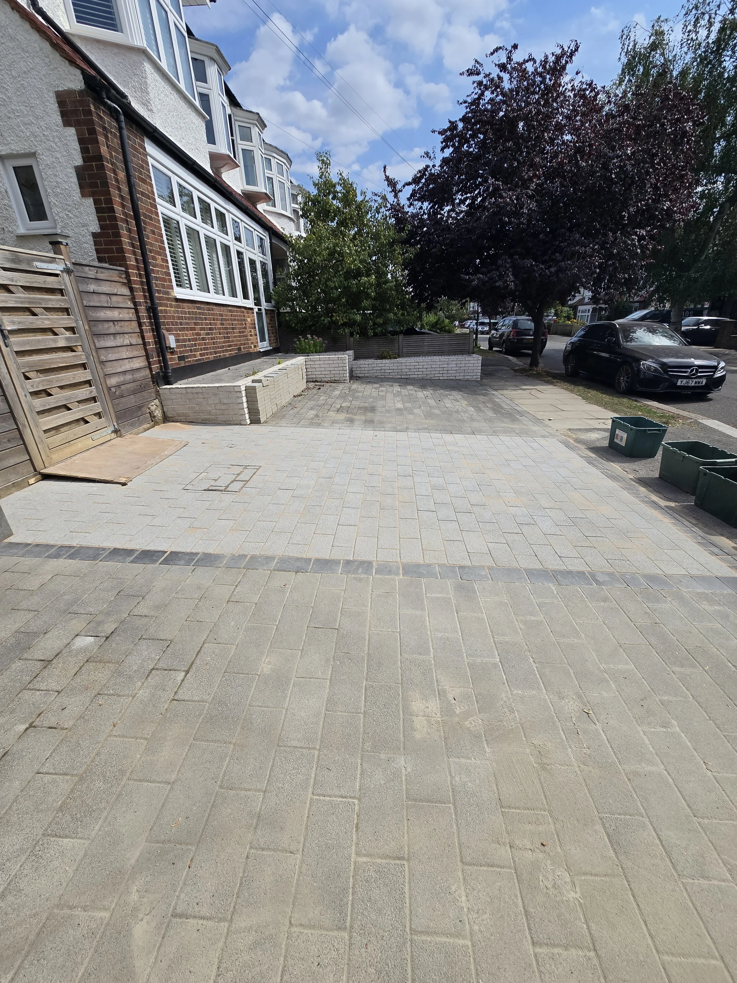 A residential front yard with a paved driveway, a wooden gate, a house with bay windows, trees, parked cars, and a partly cloudy sky.