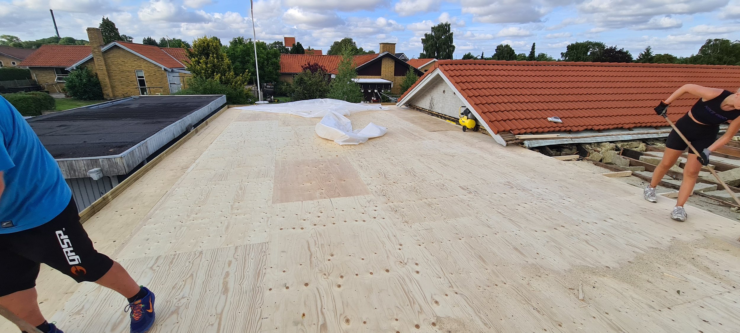 Workers are installing plywood sheathing on a flat rooftop, with neighboring houses in the background and tools visible on the roof.