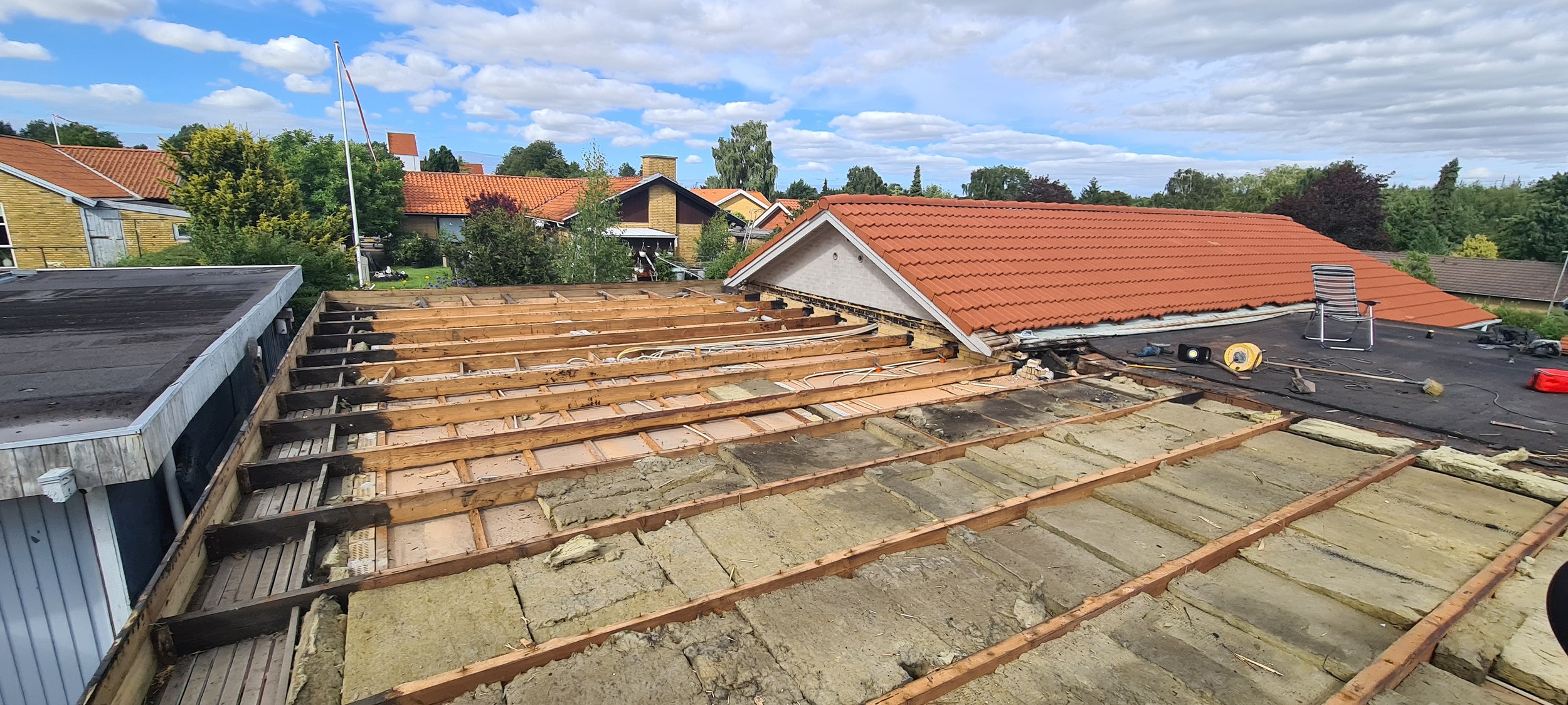 A house roof under renovation with exposed wooden framing and insulation, and neighboring houses with red tiled roofs under a partly cloudy sky.
