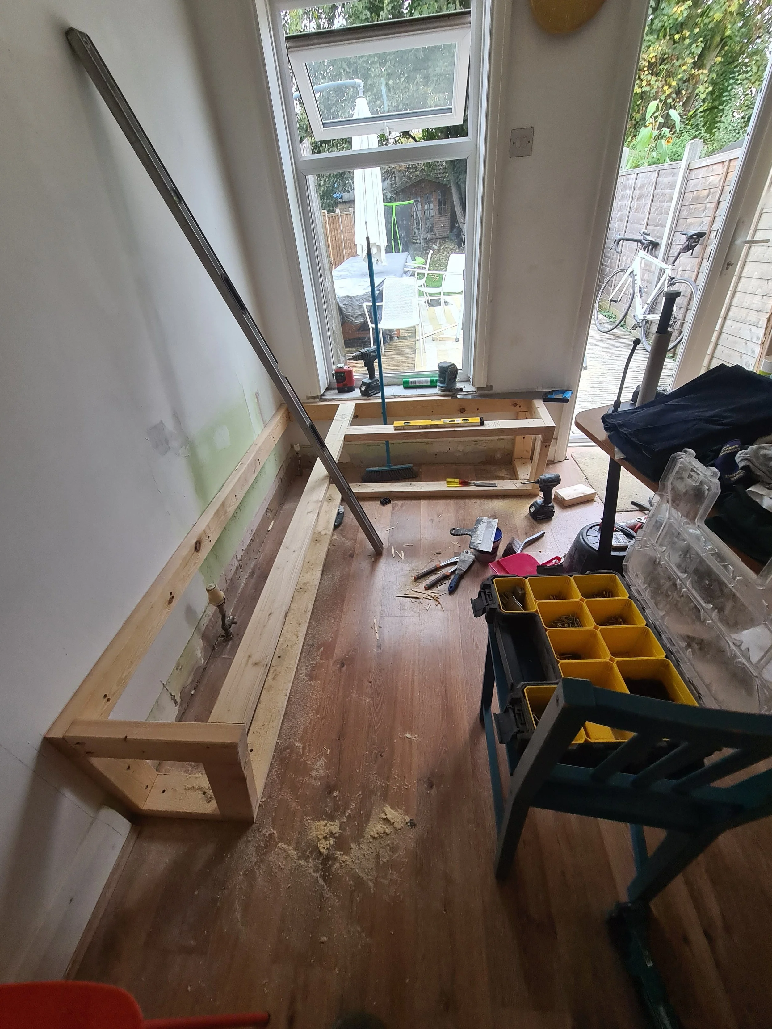 A room undergoing renovation with construction tools and materials, including a wooden frame on the floor, a level, a hammer, and a toolbox. There is a large window with an open bottom sash, a partially visible chair, and an outdoor view with a bicyc