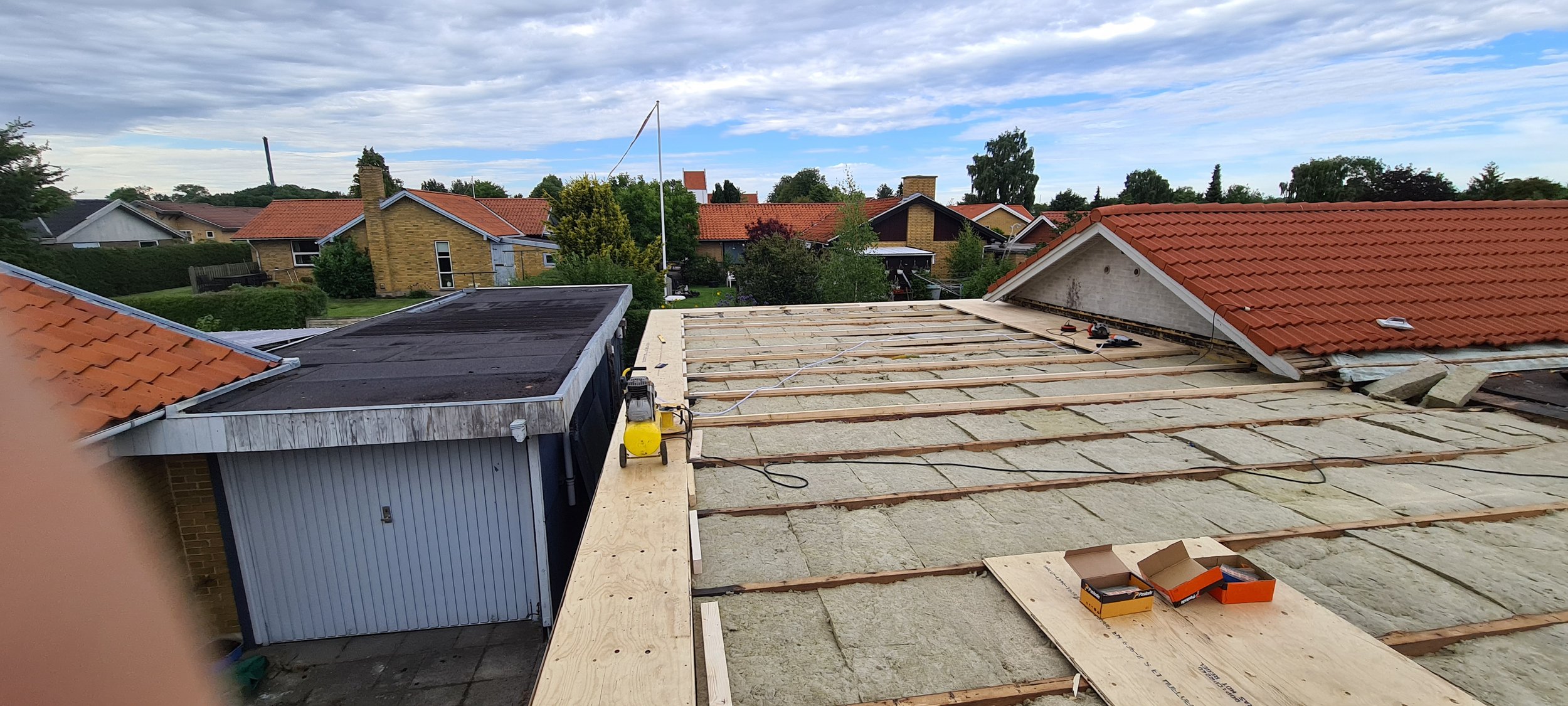 Roof under construction with exposed wooden floor and some tools, neighboring houses with red and gray tiled roofs, cloudy sky.