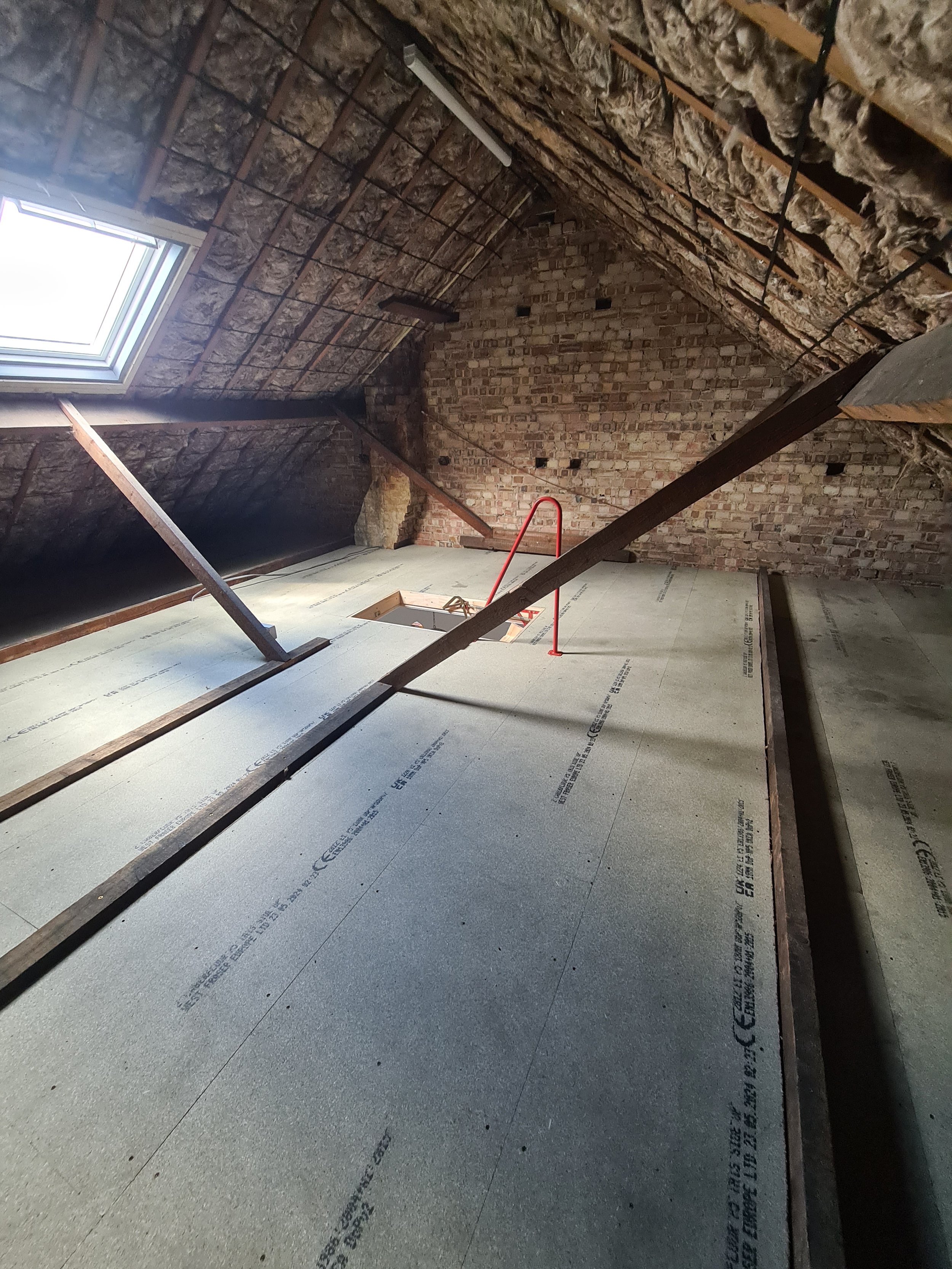 Attic with drywall installed on the floor, wooden supports and wiring, brick wall, and a skylight window on the ceiling.