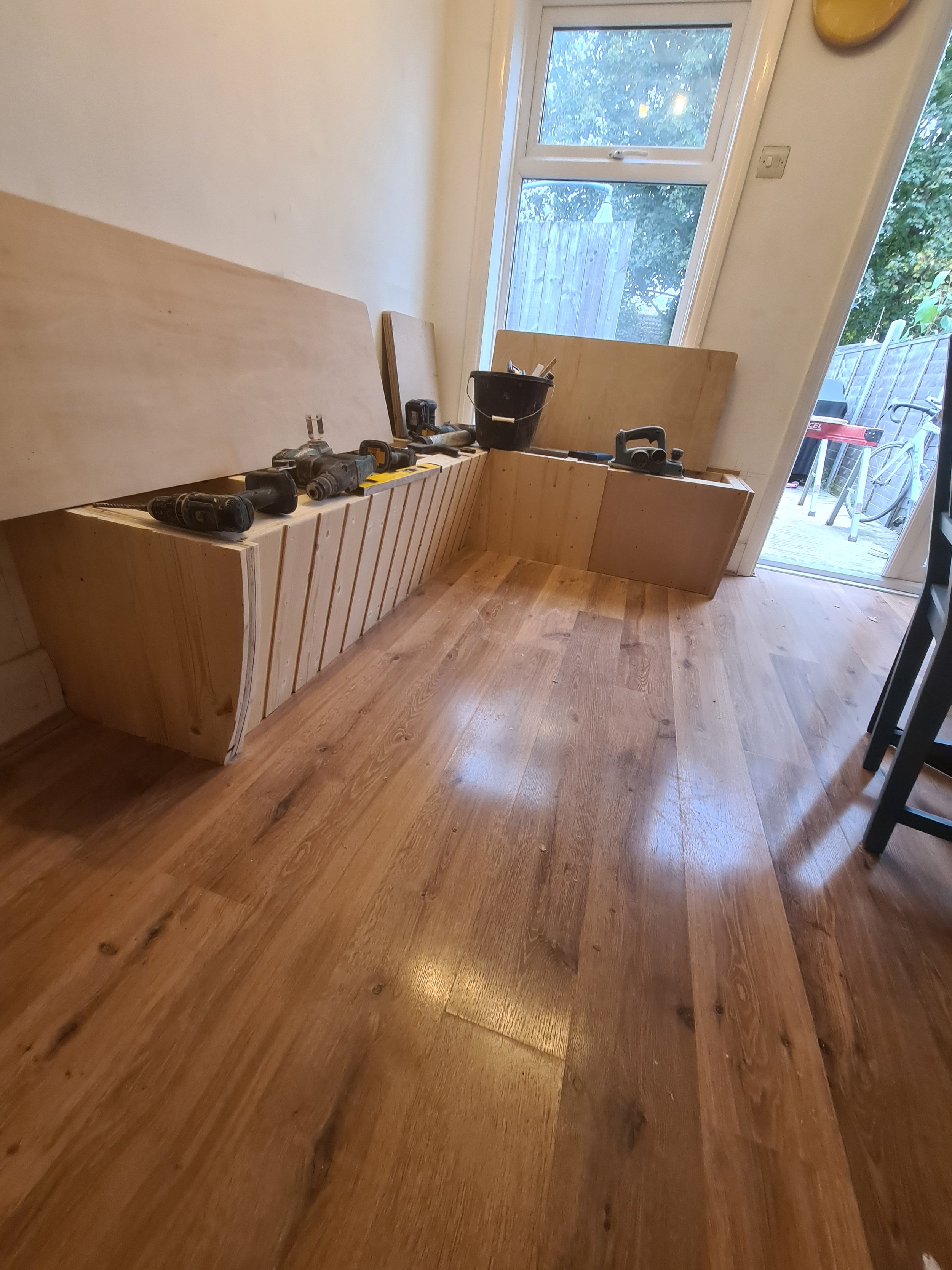 Interior of a room with wooden flooring, partially assembled wooden furniture, a black bucket, and cordless drills on the furniture, with a glass door and a window showing outdoor trees and a wooden fence.
