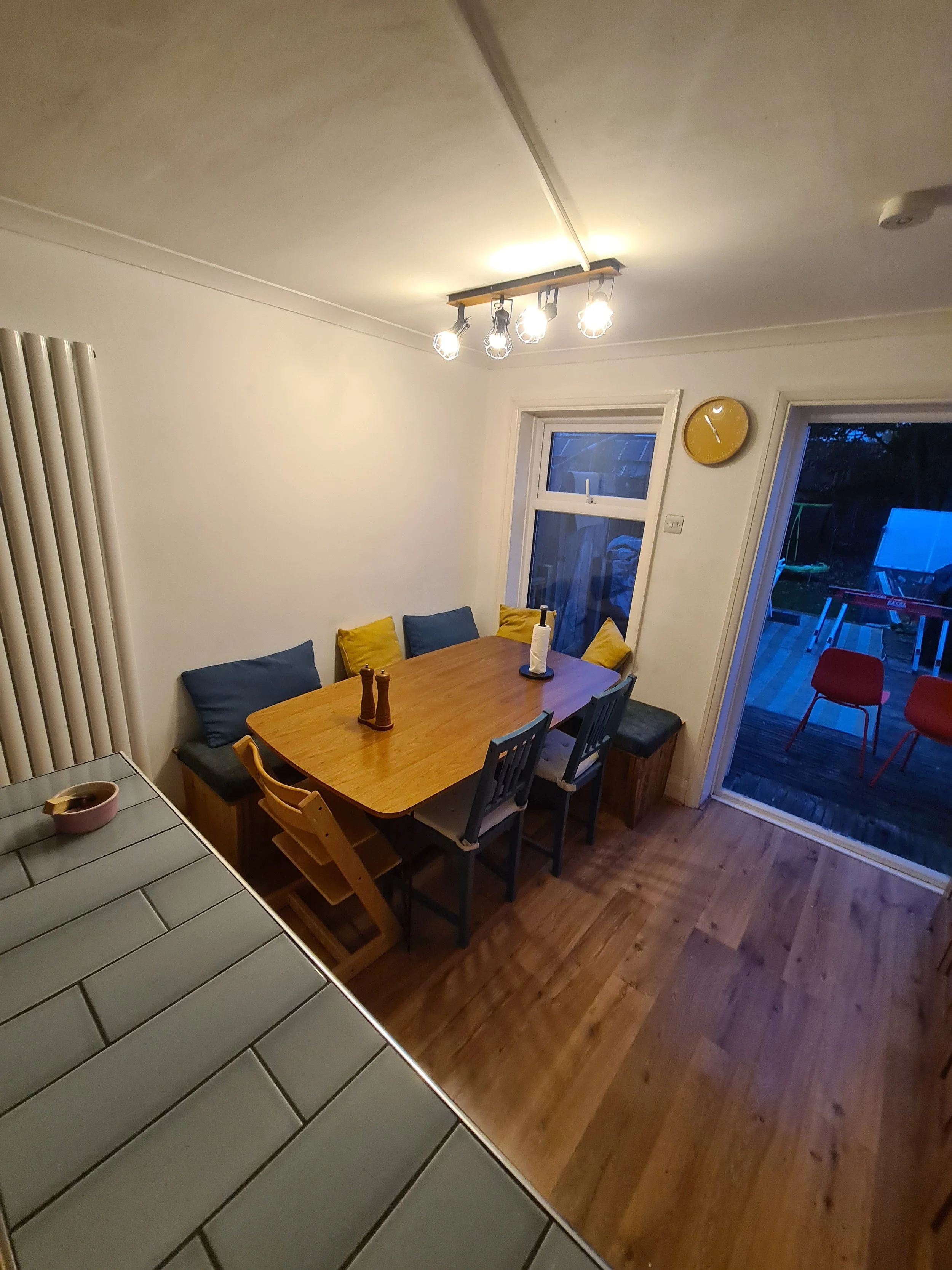 A dining area with a wooden table surrounded by six chairs, some with blue and yellow cushions. The area has two windows, a yellow wall clock, and a sliding glass door leading to an outdoor deck with red chairs and a swing set. Overhead, there is a c