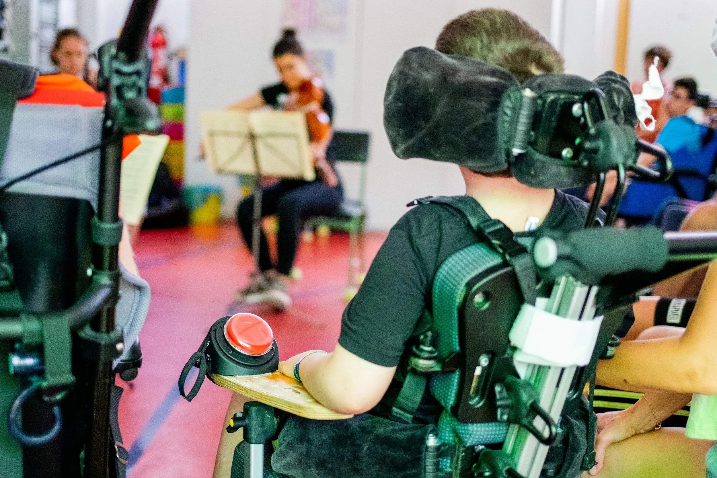 Niño en silla de ruedas en un aula, rodeado de otros niños, una niña tocando el violín en el fondo y otros niños en diversas actividades.