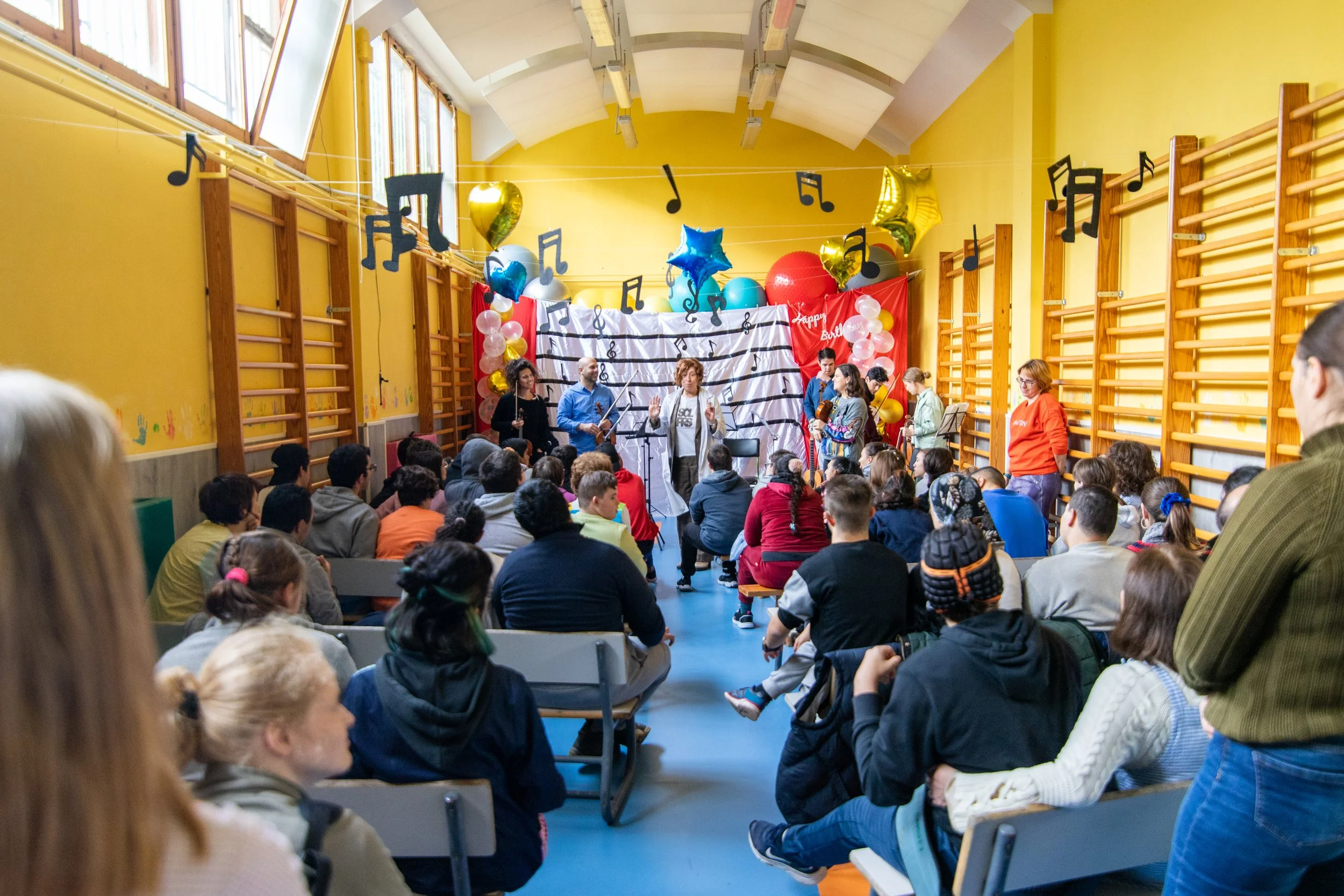 Niños y adultos en un salón de escuela durante una actuación o celebración, decorado con globos, dibujos en las paredes y notas musicales colgando en el aire.