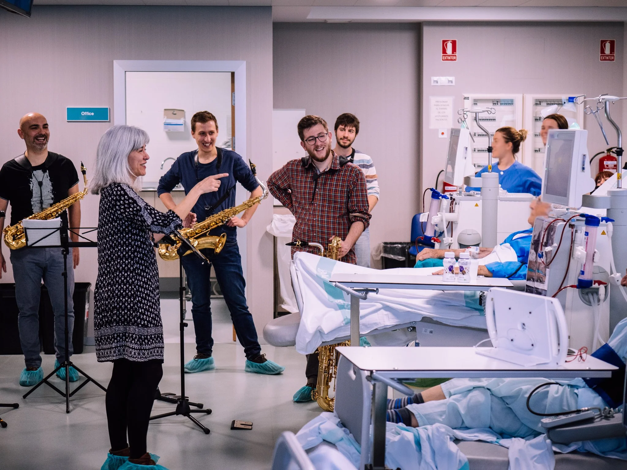 Un grupo de personas tocando saxofones en un hospital, mientras varios pacientes están en sus camas dentro de la sala de hospital, algunos con tubos y monitores.