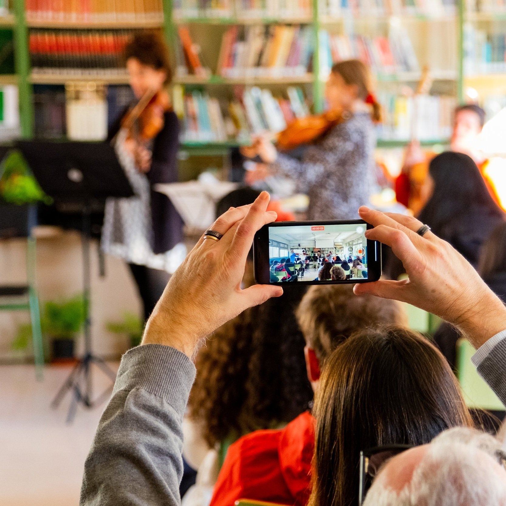 Persona grabando una orquesta tocando en una sala de clases o biblioteca con su teléfono móvil.