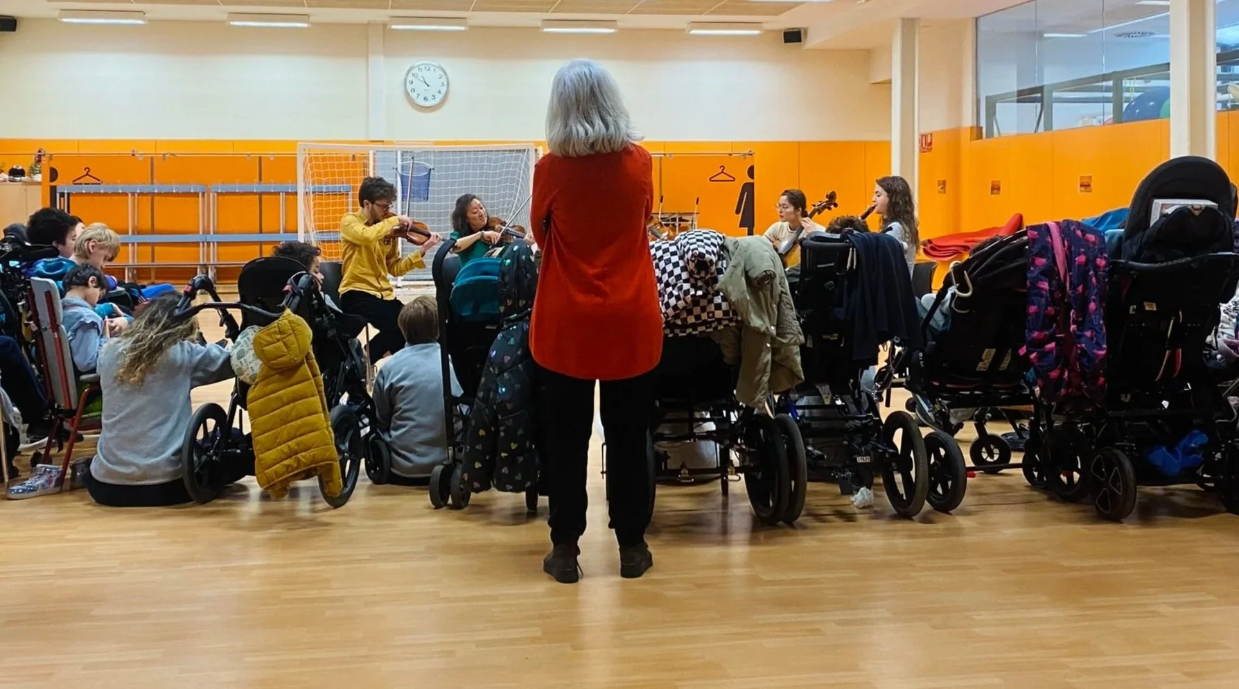 Un grupo de niños en una sala de ejercicios o actividades, algunos en sillas de ruedas, observando una orquesta de adultos tocando violín, con un reloj en la pared y paredes naranjas.