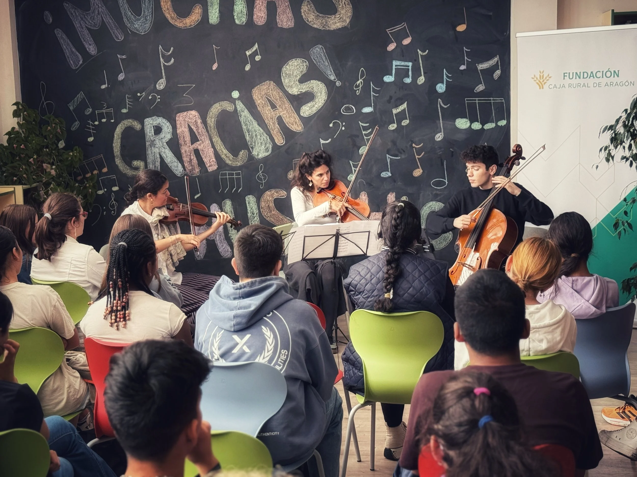 Grupo de músicos tocando violín y violonchelo frente a una audiencia en un evento con una pizarra de fondo decorada con notas musicales y la palabra 'Gracias!'.