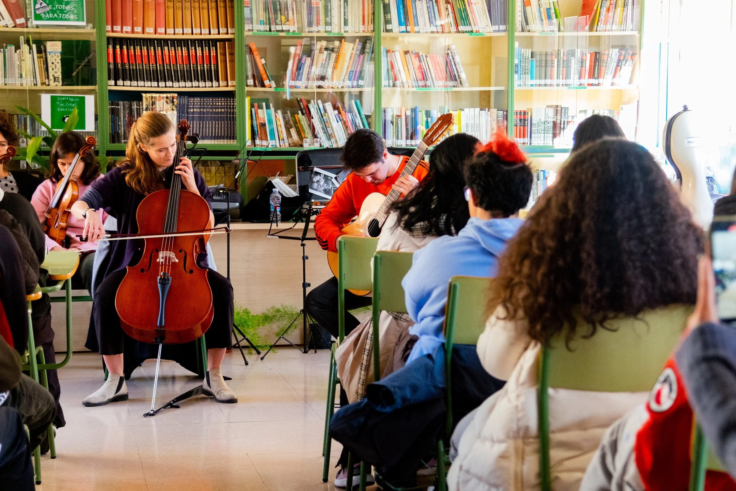 Niños y jóvenes tocando instrumentos de cuerda en una biblioteca, rodeados de libros en estantes y un grupo de personas viendo la presentación.