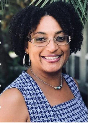 Smiling woman with glasses, hoop earrings, and a necklace, wearing a sleeveless blue patterned top, outdoors with greenery in the background.