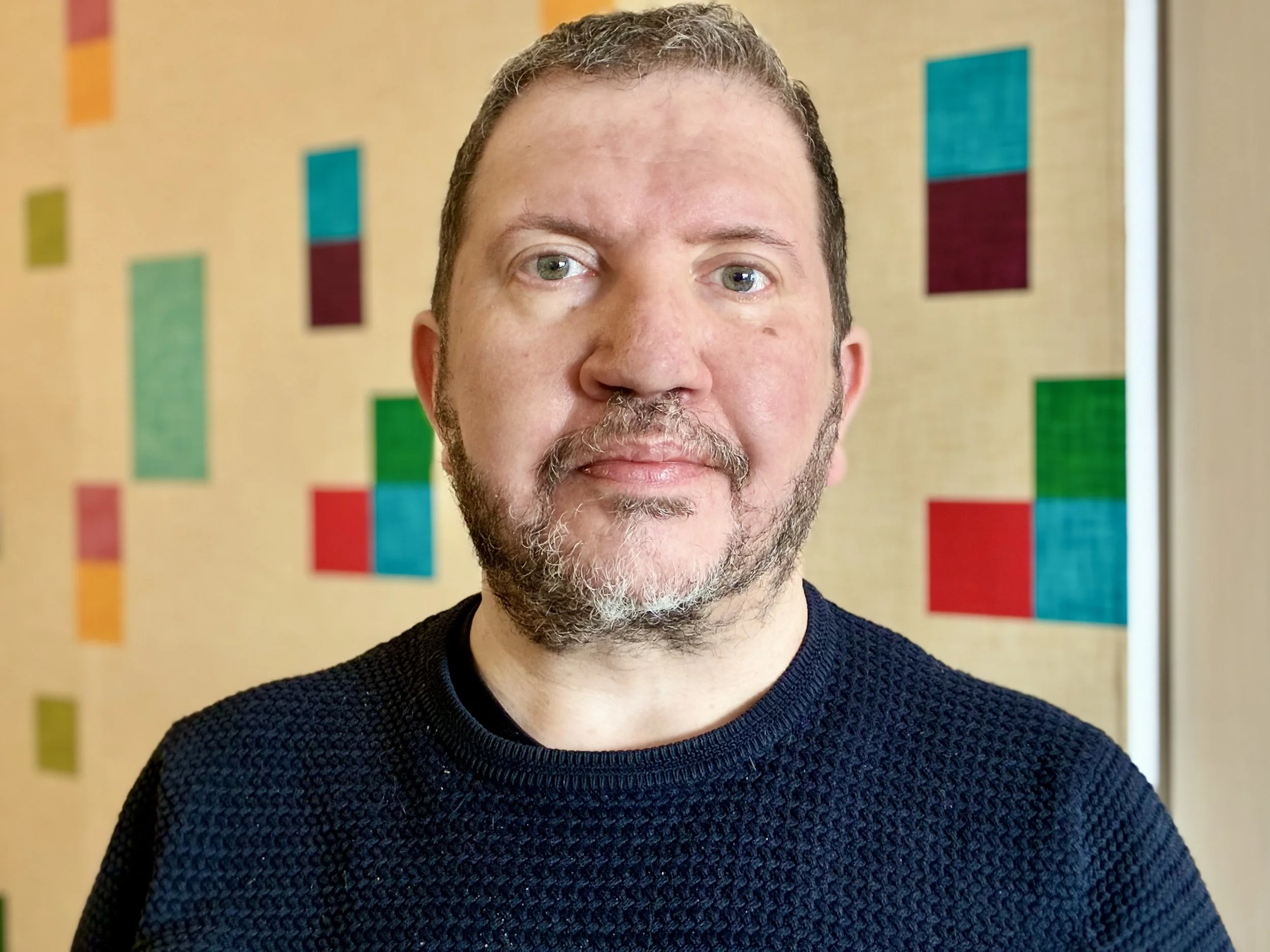 Un homme avec une barbe brune et un pull noir devant un mur avec des blocs colorés.