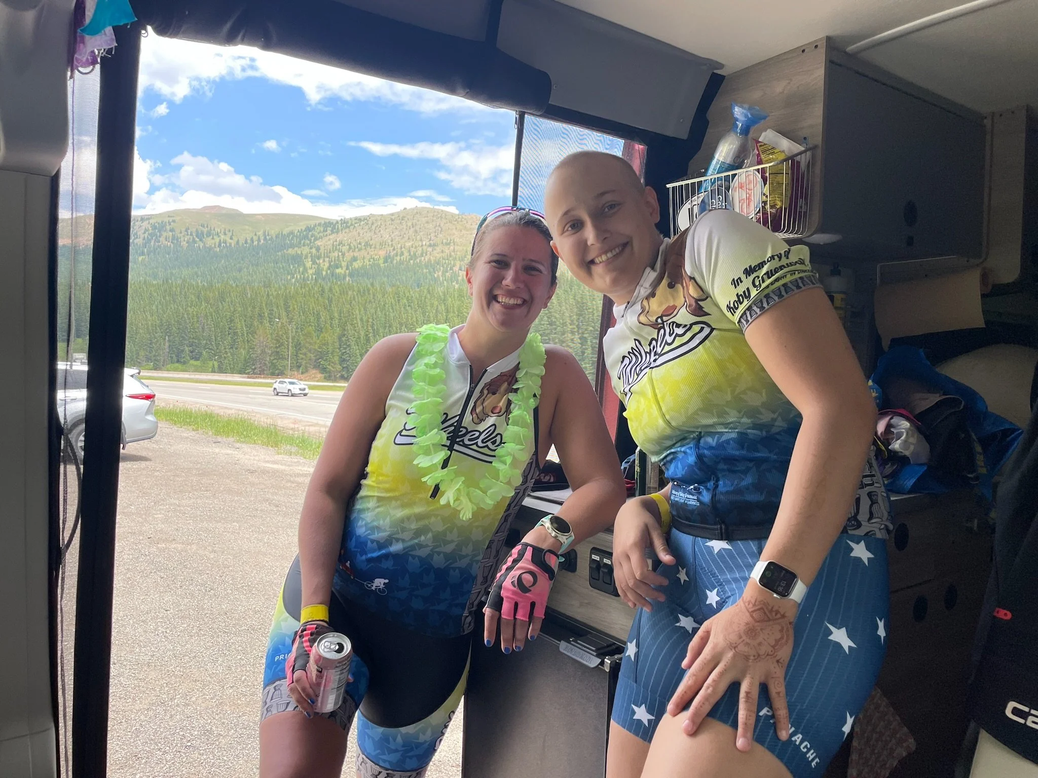 Two women in cycling gear smiling and posing inside a vehicle with a scenic mountain and forest view outside.
