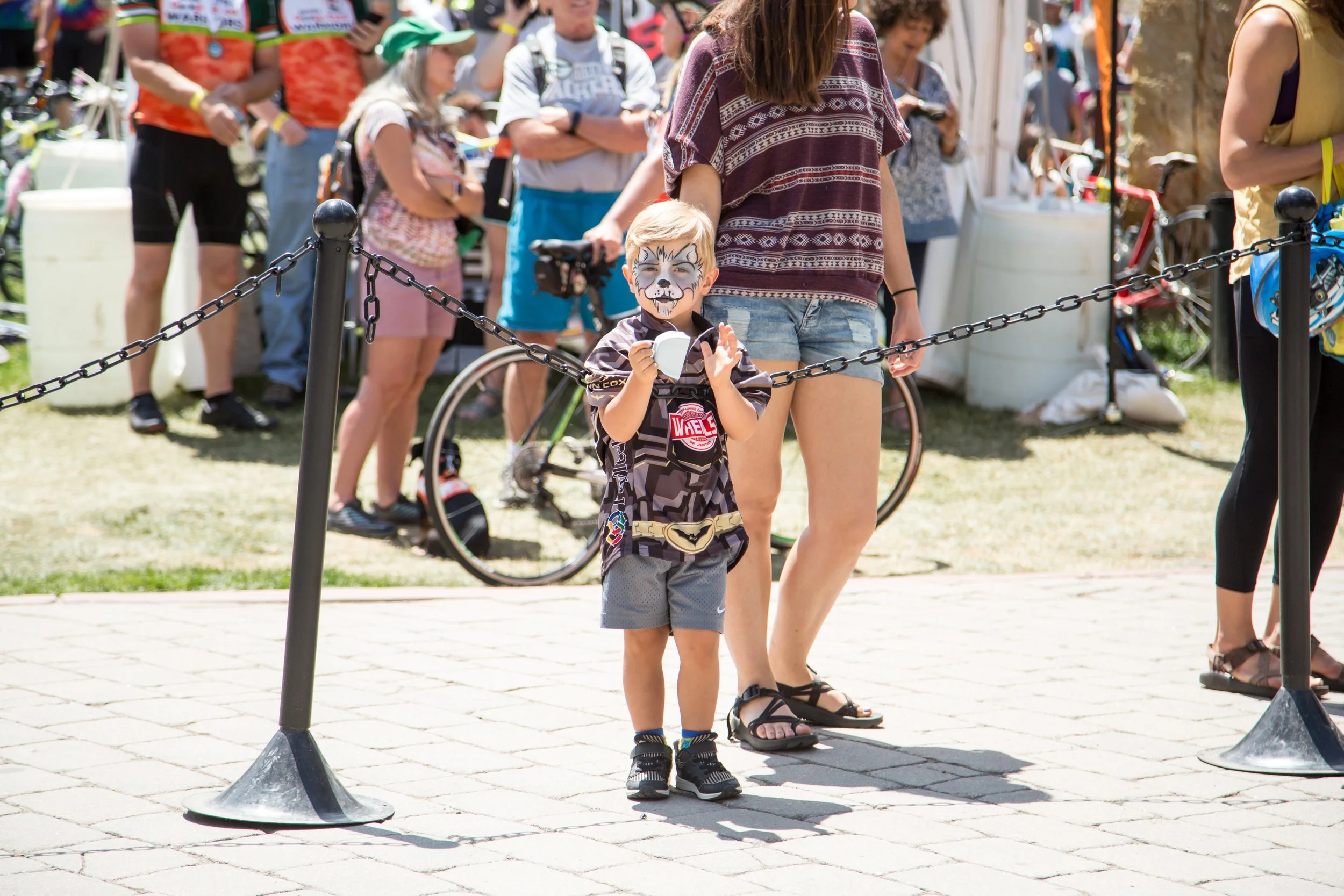 A young boy with face paint resembling a wolf, wearing sports clothes, stands behind a black chain barrier at an outdoor event, holding a small object in his hands. Behind him, a woman in shorts and a patterned shirt is also near the barrier. There a
