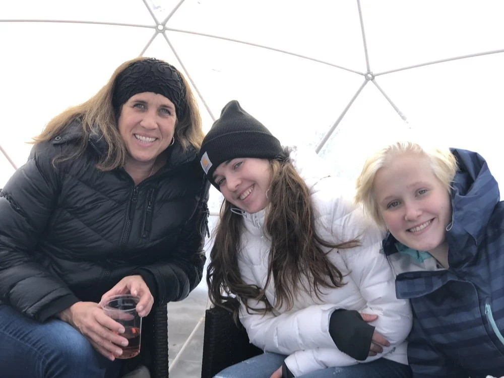 Three women sitting closely together inside a geodesic dome, smiling at the camera, dressed in winter clothing and hats.