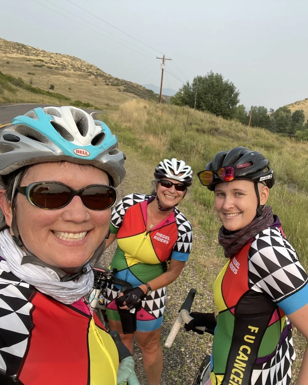 Three smiling women in cycling gear and helmets taking a selfie outdoors on a rural road with hills and trees in the background.