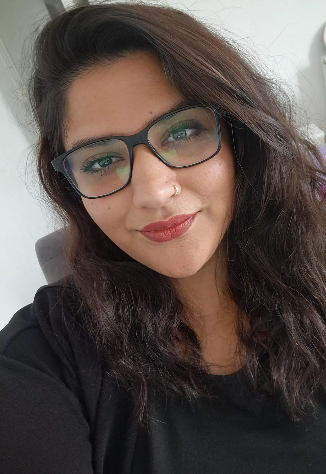 A woman with shoulder-length wavy dark brown hair, wearing black glasses, a gold nose ring, and a black top, smiling at the camera.