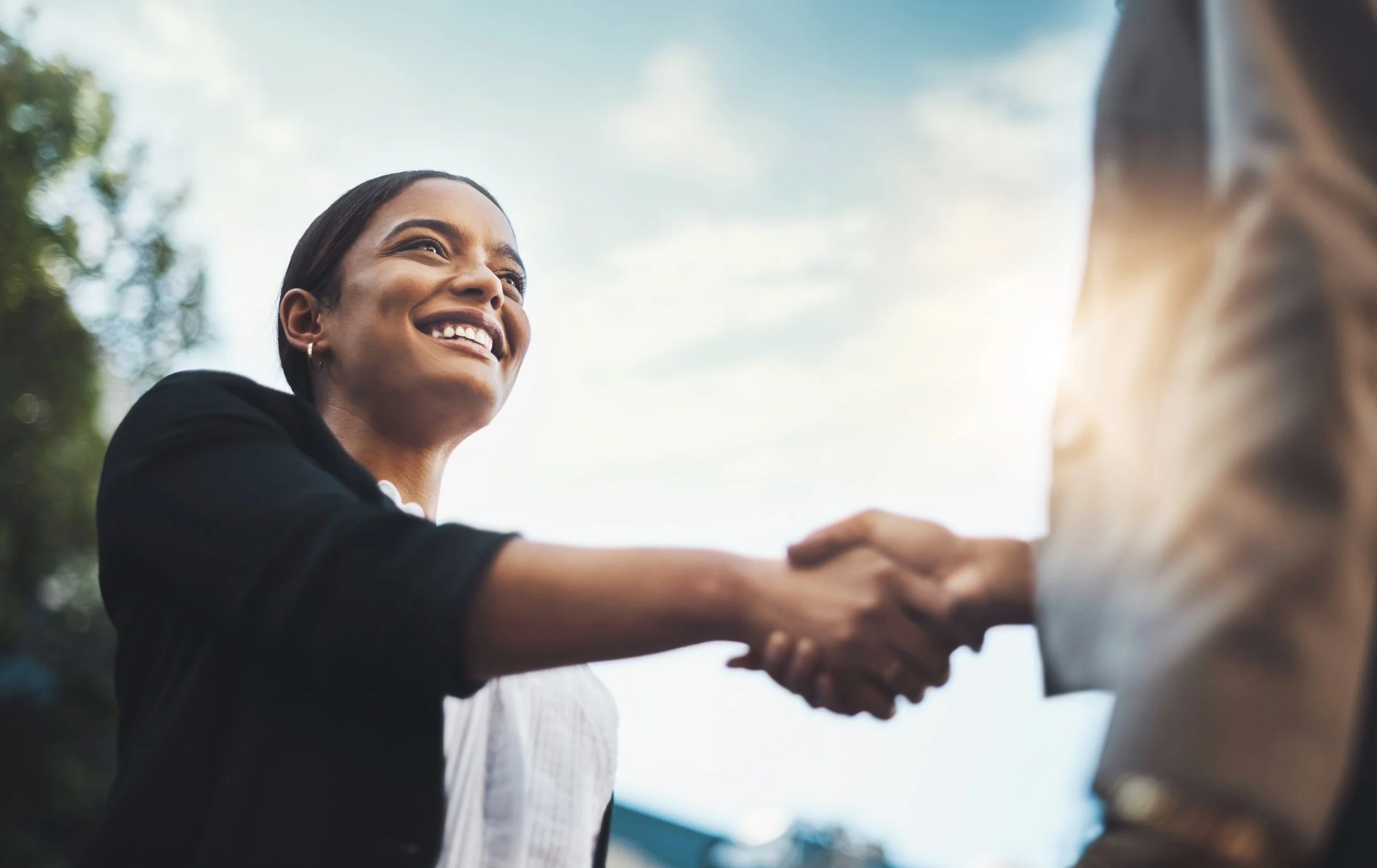 A woman smiling and shaking hands with someone outdoors under a bright sky.