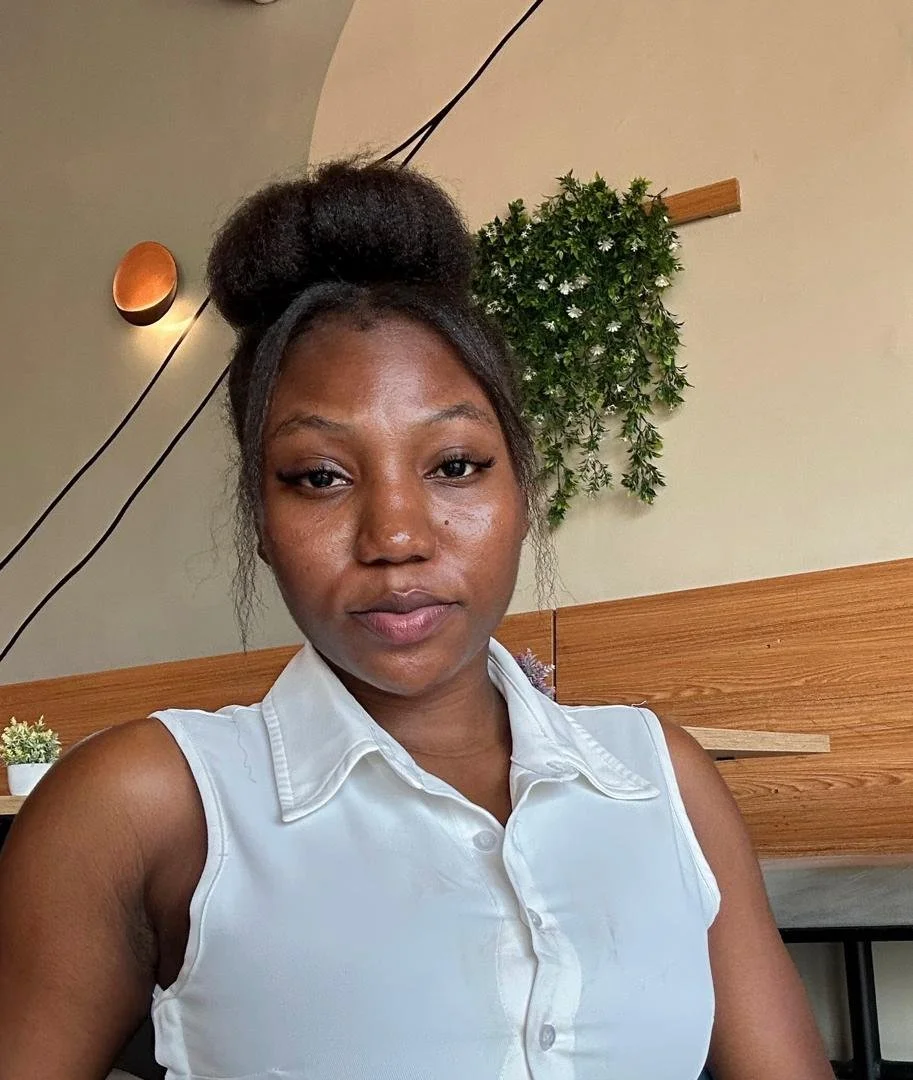 A young woman with dark hair in a bun, wearing a white sleeveless collared shirt, sitting indoors with a wooden wall and green plant in the background.