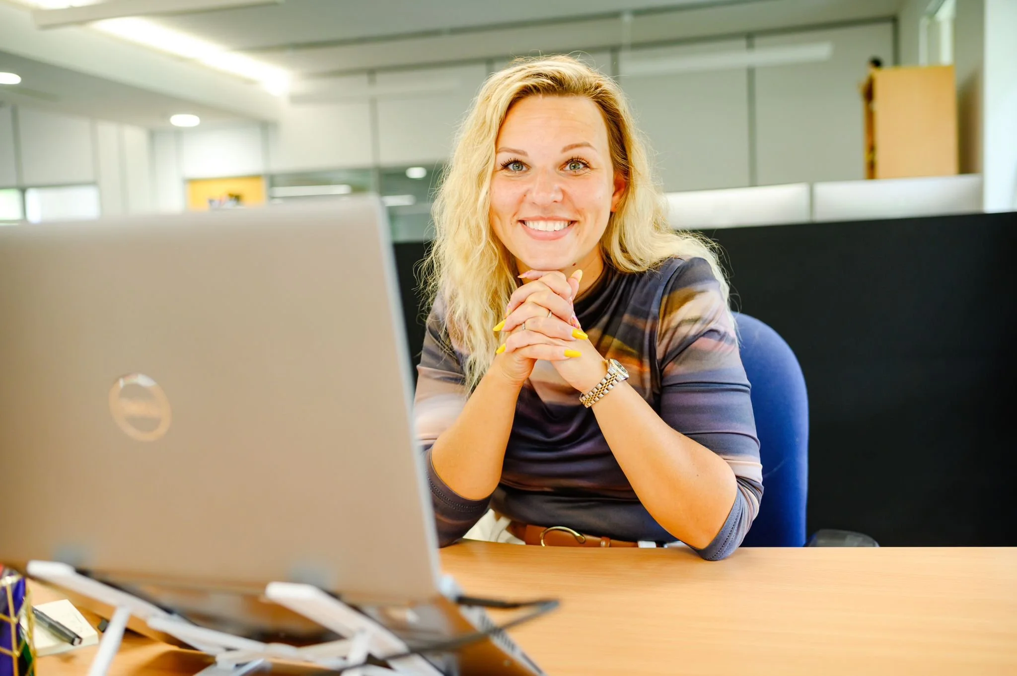 A woman with blonde curly hair sitting at a desk in an office, smiling with her hands clasped together in front of her. There is a laptop on the desk and the office has a modern appearance with cubicles and glass walls.