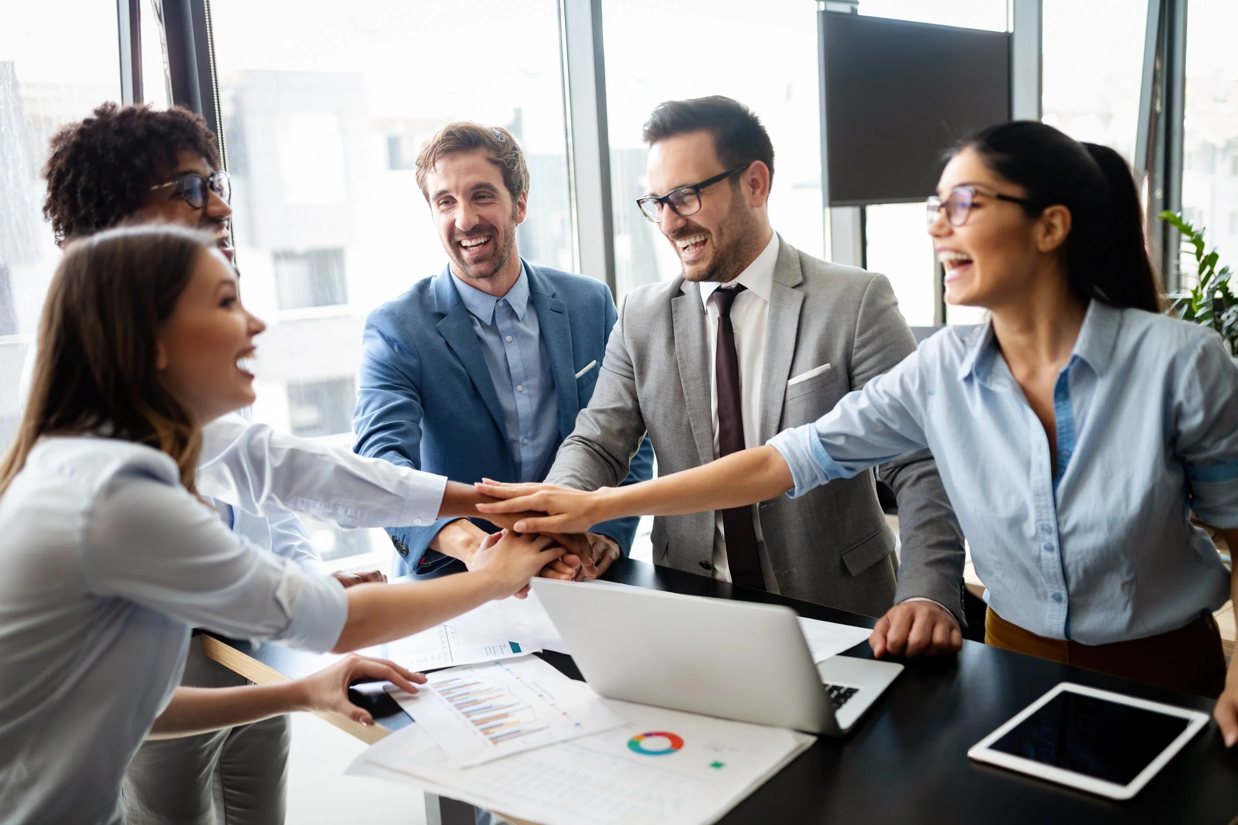 Five diverse professionals in business attire smiling and placing hands together in a team huddle in a modern office with large windows.