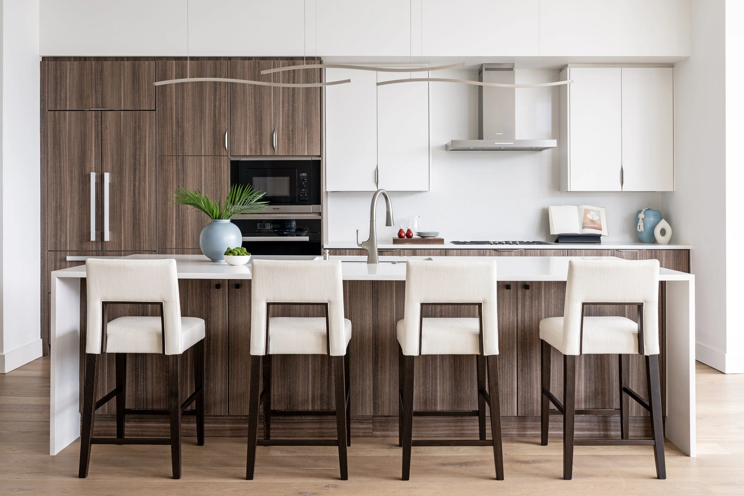 Modern kitchen with wooden cabinetry, white countertops, four beige upholstered bar stools, and decorative vases on the countertop.