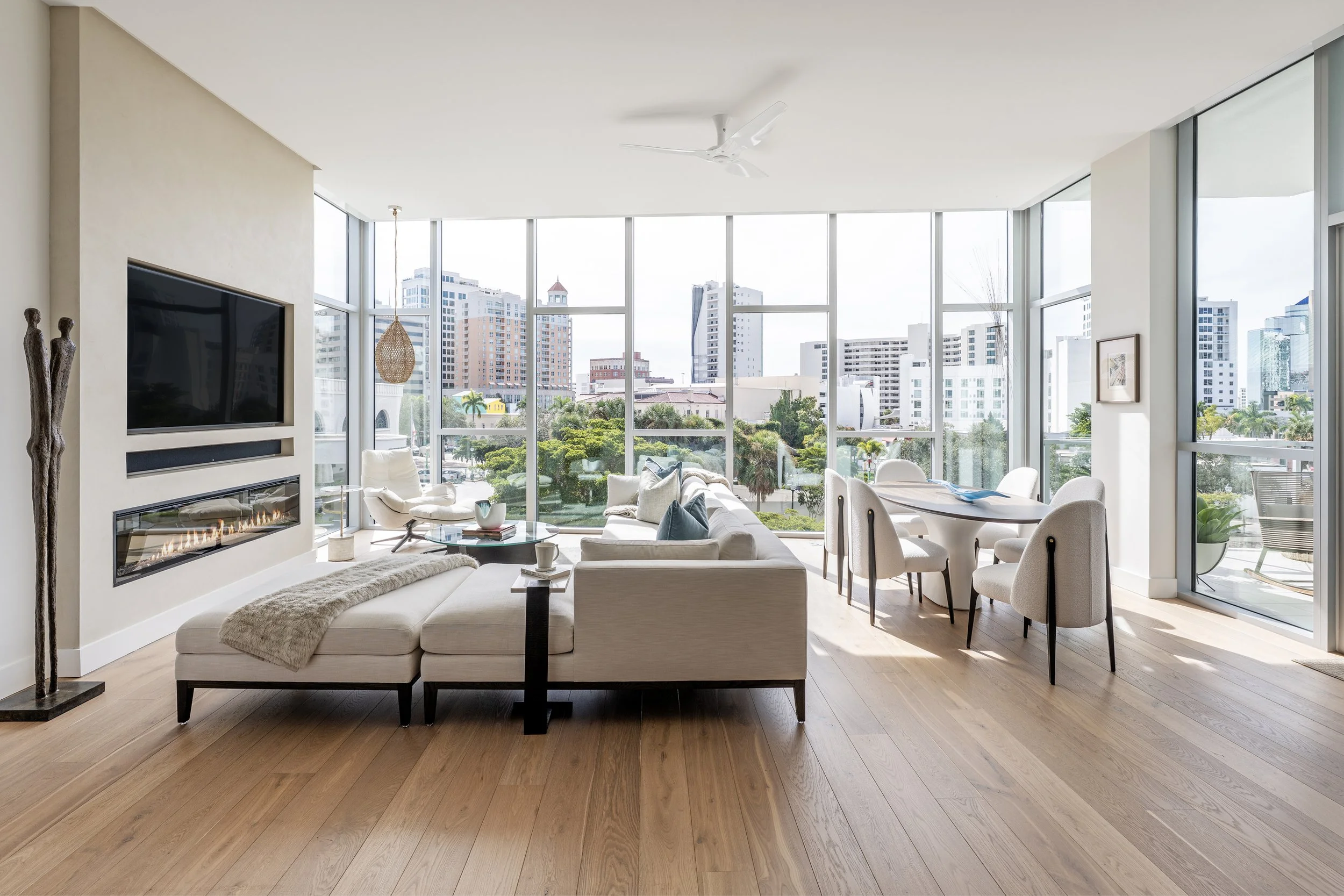 Modern living room with large windows, white sofa, coffee table, dining table with chairs, and cityscape view