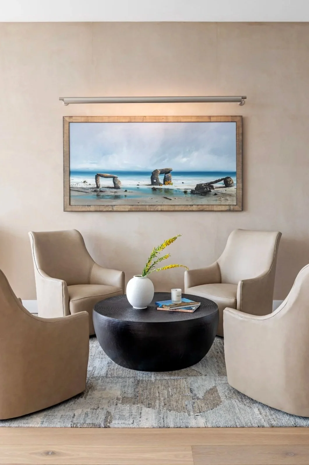 A cozy living room with four beige armchairs arranged around a black circular coffee table. On the coffee table, there is a white vase with yellow and green flowers, a small white candle, and some magazines. A large framed painting of Stonehenge on a beach hangs on the wall behind the chairs, and a modern silver picture light is mounted above the painting.
