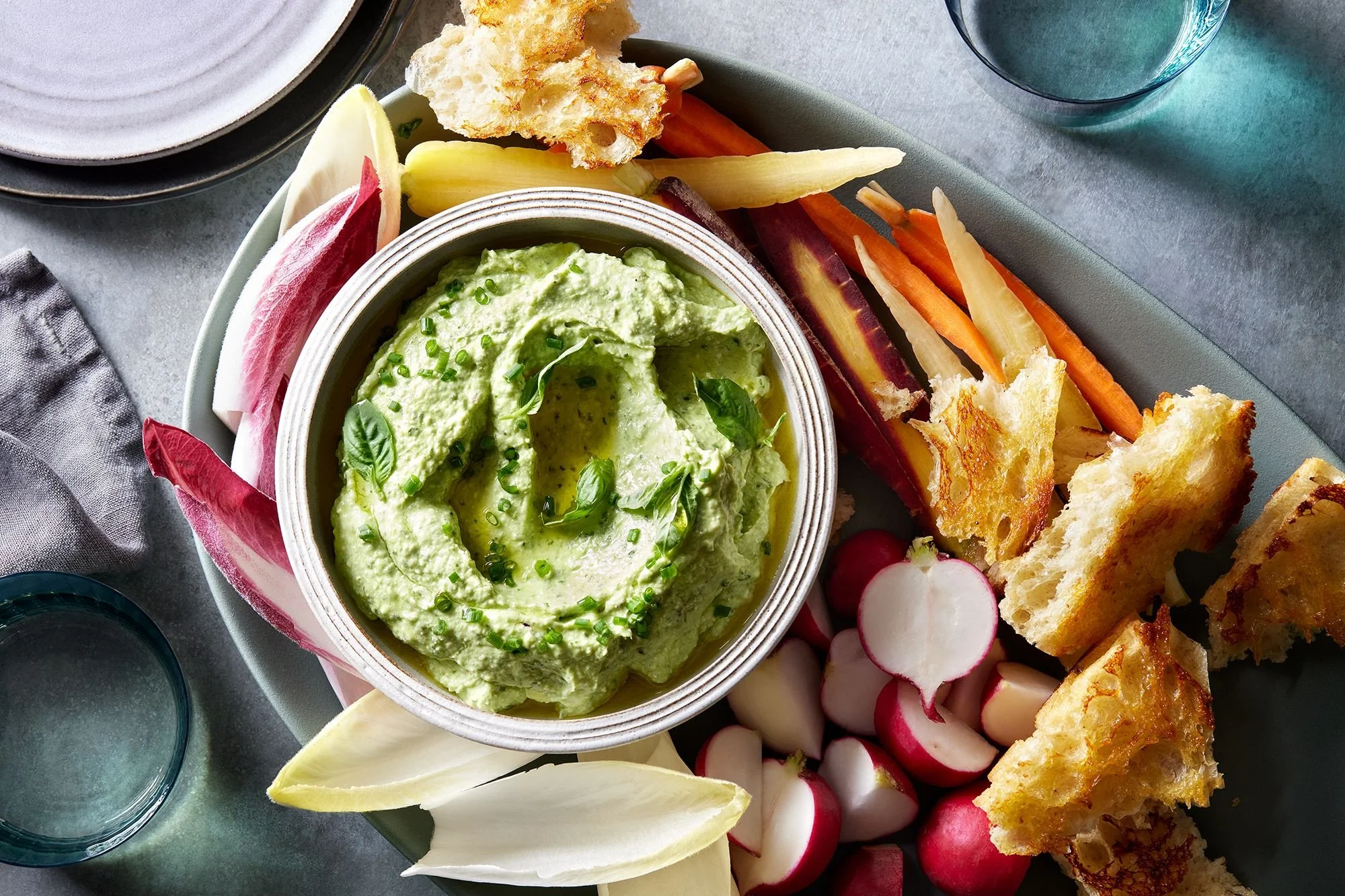 A plate of assorted vegetables and pita chips served with herbed feta. The vegetables include radishes, carrots, parsnips, and sliced onions, with some artichoke hearts. The pita chips are crispy and golden.
